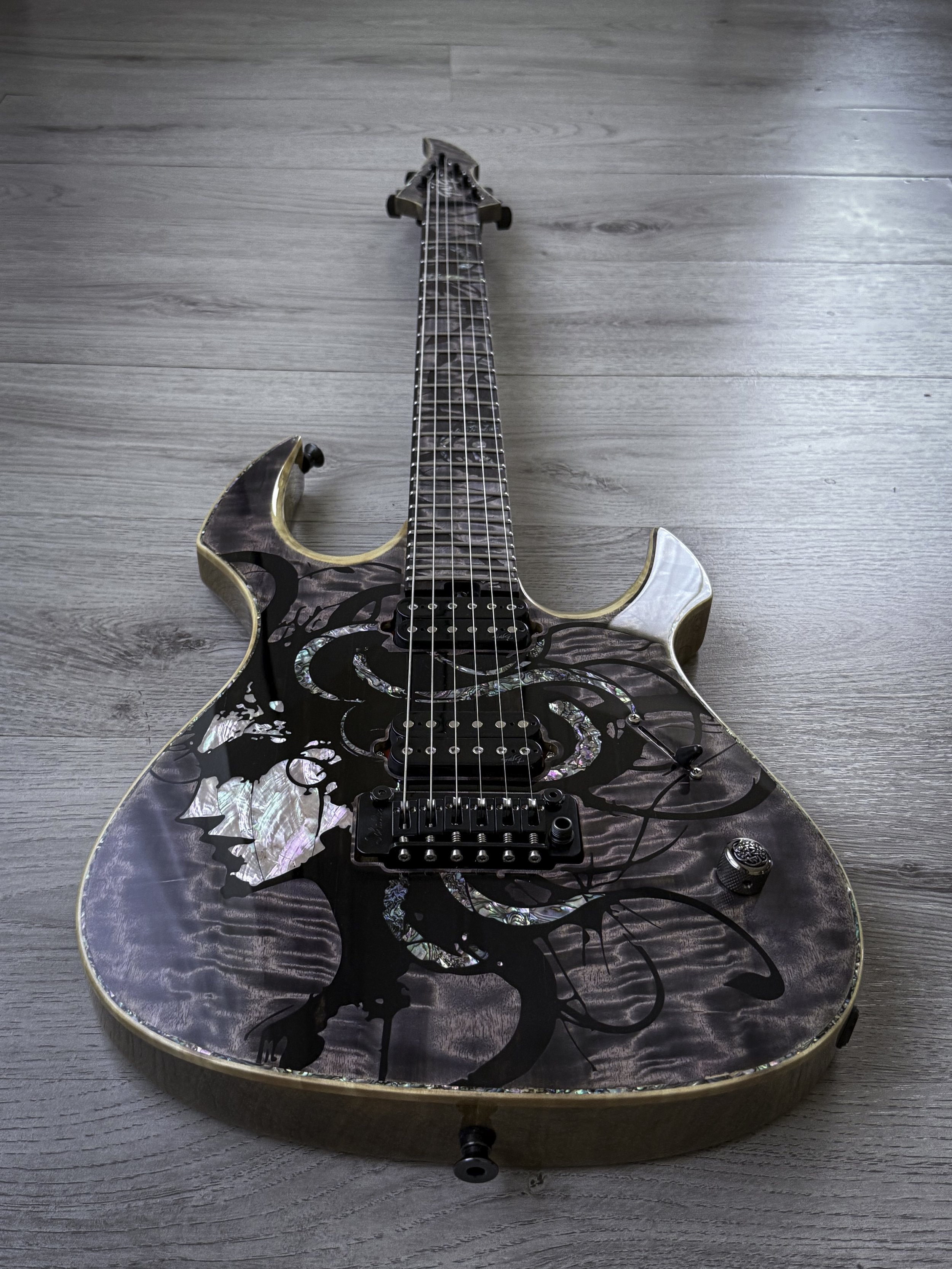 GNG Giulio Negrini electric guitar with a dark, intricate artistic design featuring abstract shapes and iridescent shell accents, lying on a wooden floor.