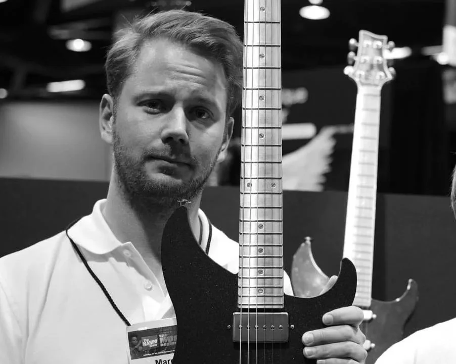 Marcel Fehse, a man with short hair and a beard holding a black electric guitar at a music event, with other guitars visible in the background.