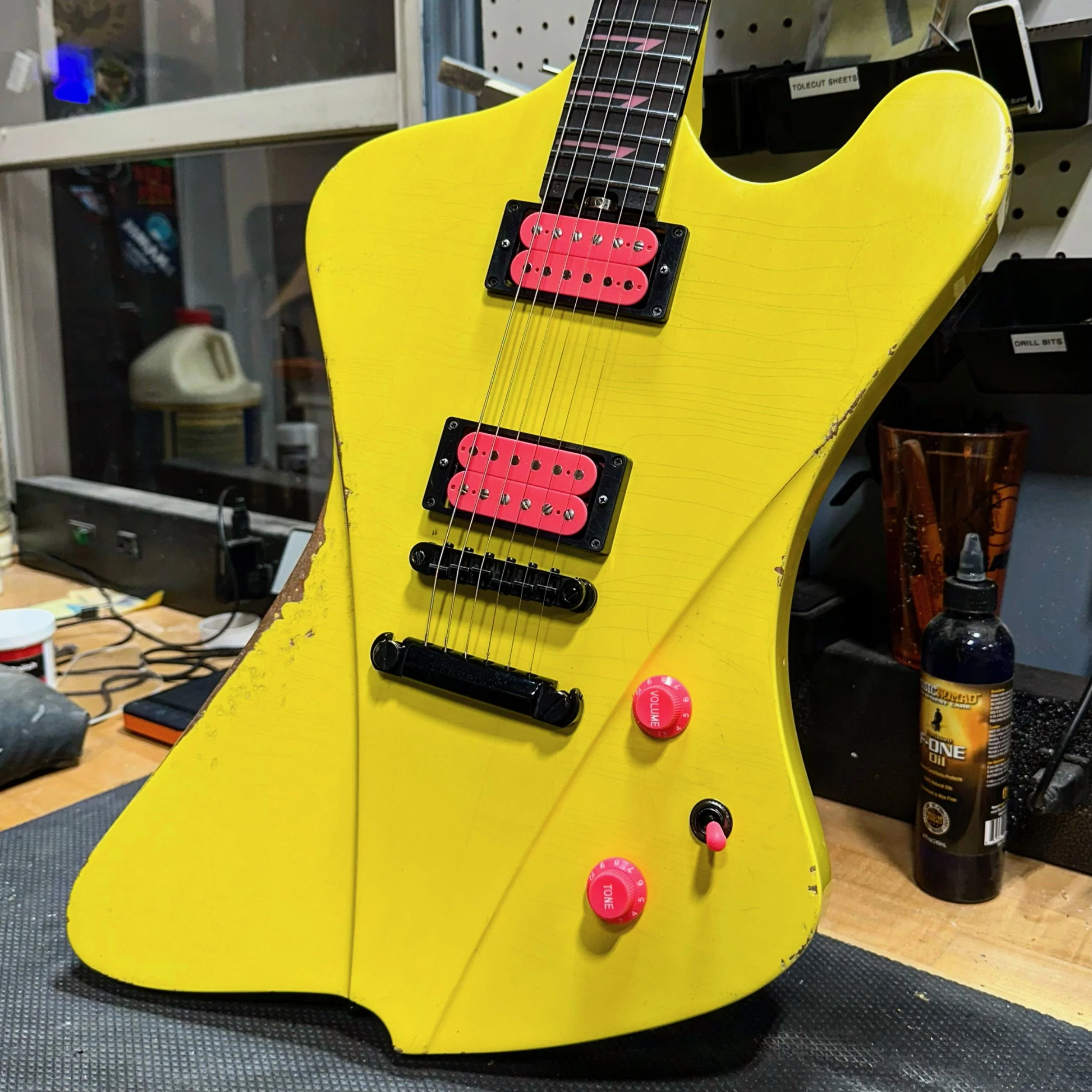 A yellow electric Sully guitar with pink pickups and control knobs, resting on a workbench in a workshop, with various tools and supplies in the background.