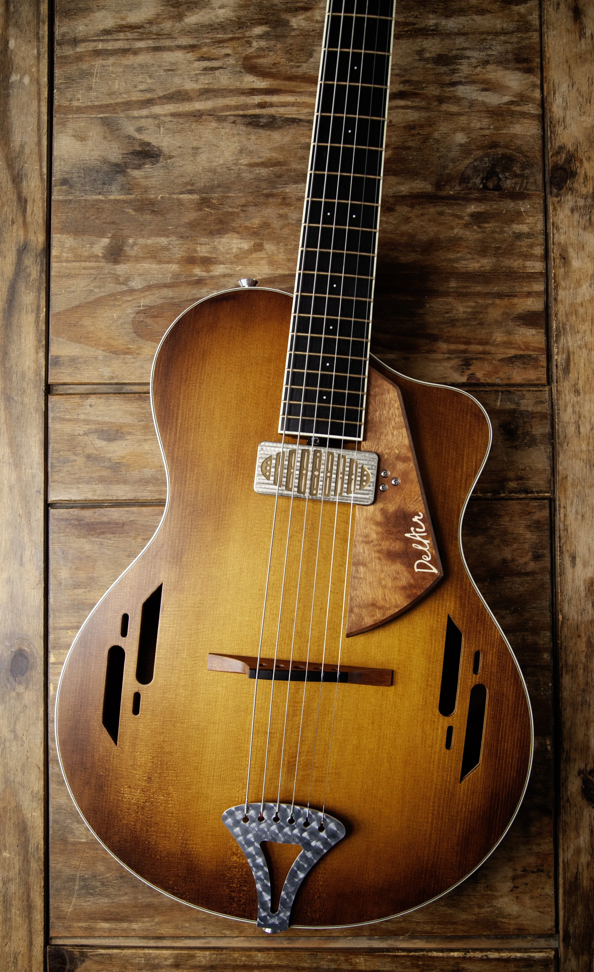 A brown acoustic electric Island Instrument guitar with a cutaway body shape, multiple sound holes, and a decorative pickguard, resting on a wooden surface.