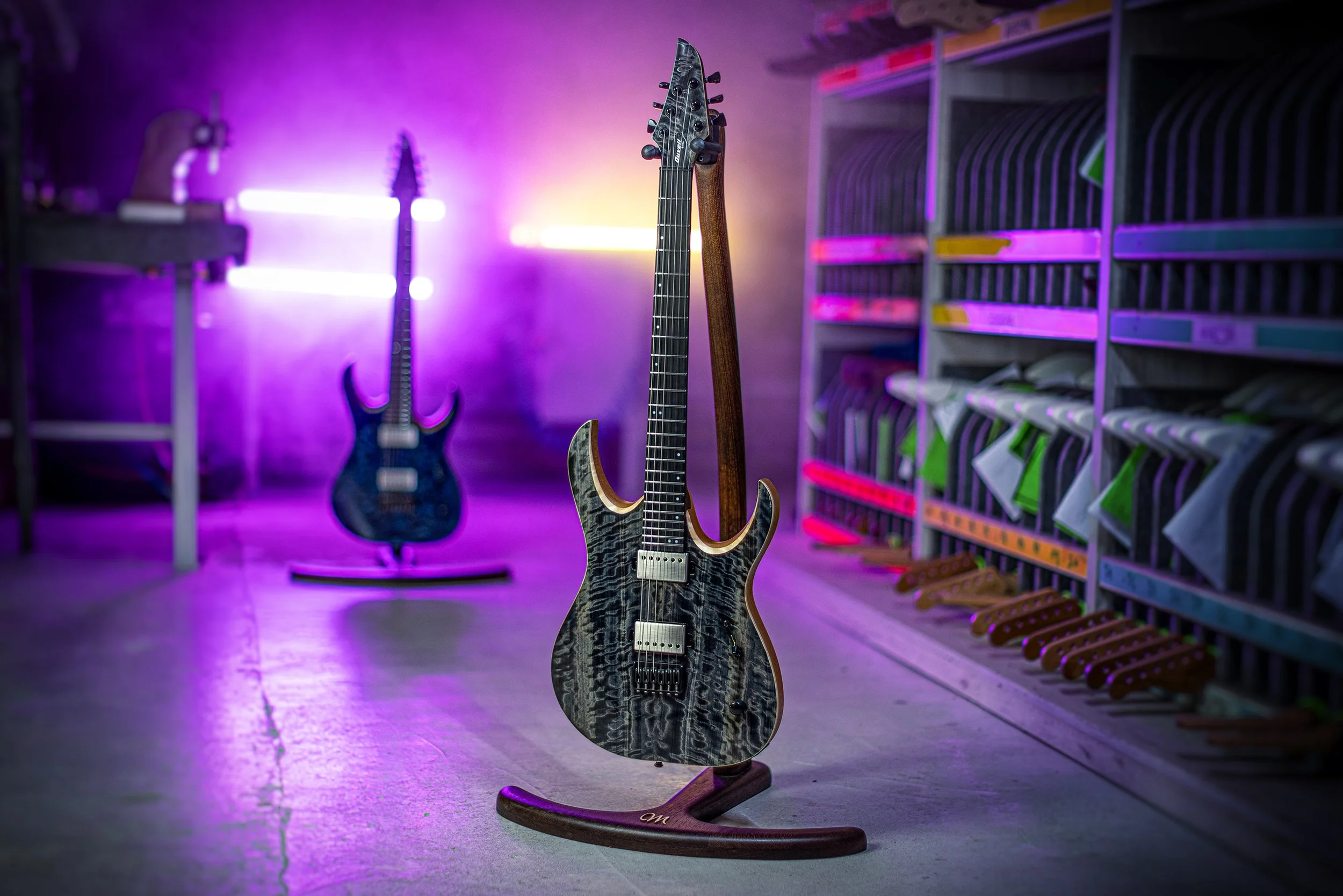 Two electric Mayones guitars, one in the foreground on a stand, featuring a quilted black and gray wood grain pattern, and one in the background leaning against a wall, both in a room with multicolored neon lighting.