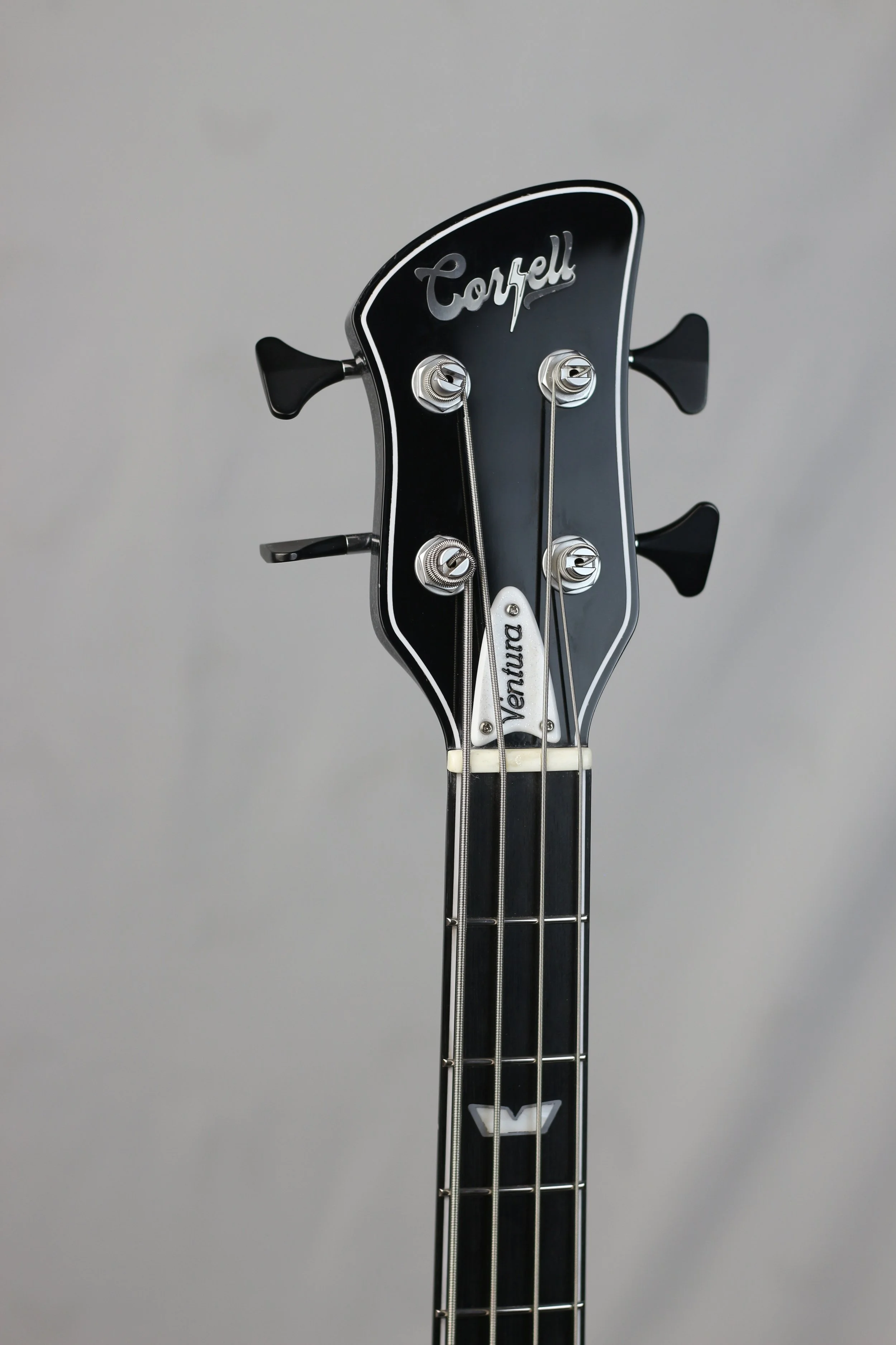 Close-up of a black bass guitar headstock with tuning pegs, strings, and the brand name "Corsell" on top, and a white "Ventura" label.