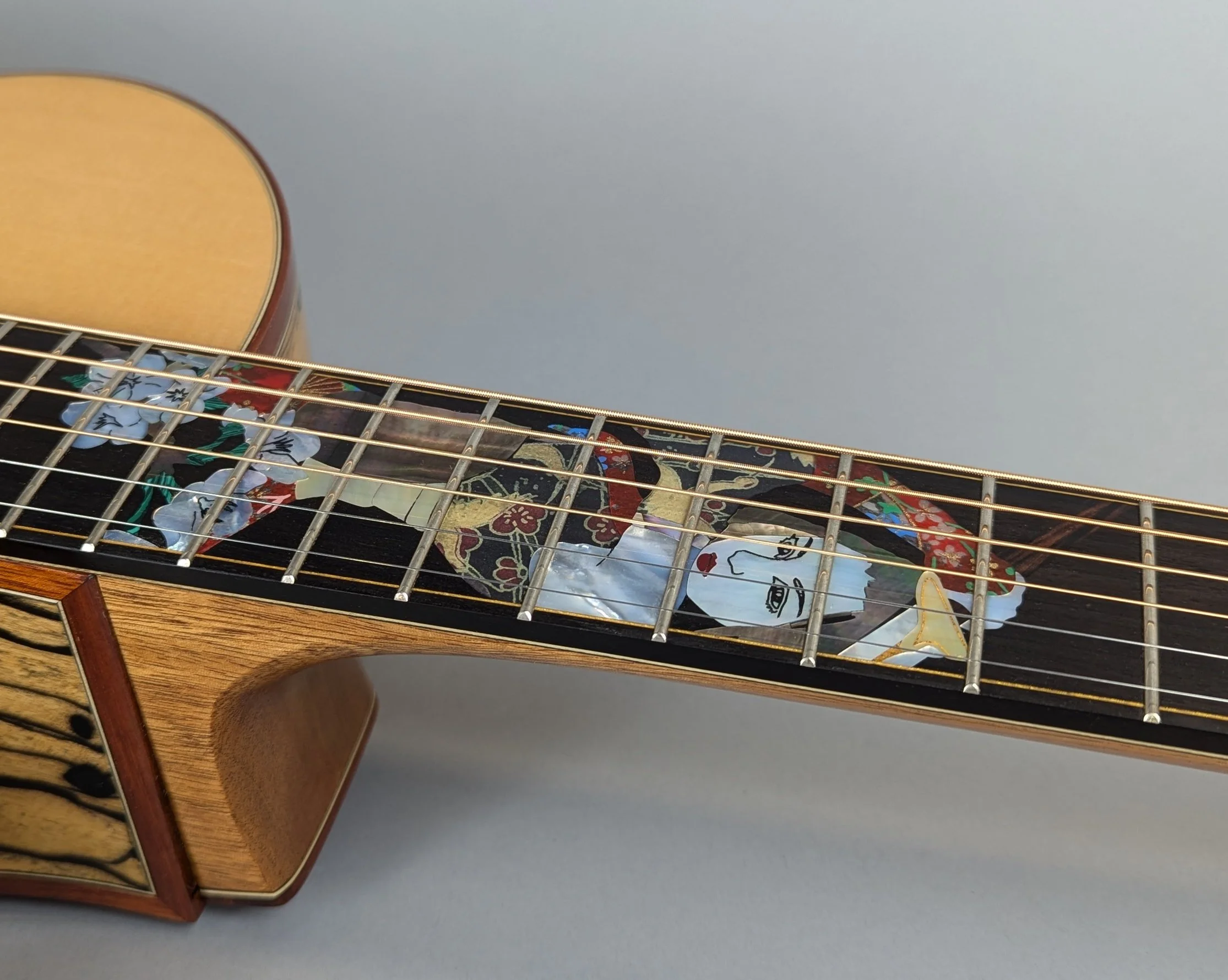 Close-up of a Morgan Kelsey guitar with a decorative inlay on the fretboard featuring a geisha design with flowing hair and floral patterns, against a plain gray background.
