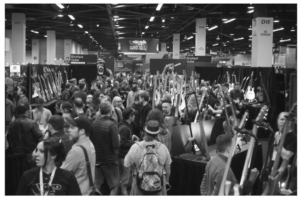 Crowd of people at the NAMM Show Boutique Guitar Showcase, browsing and viewing guitars displayed on tables and stands.
