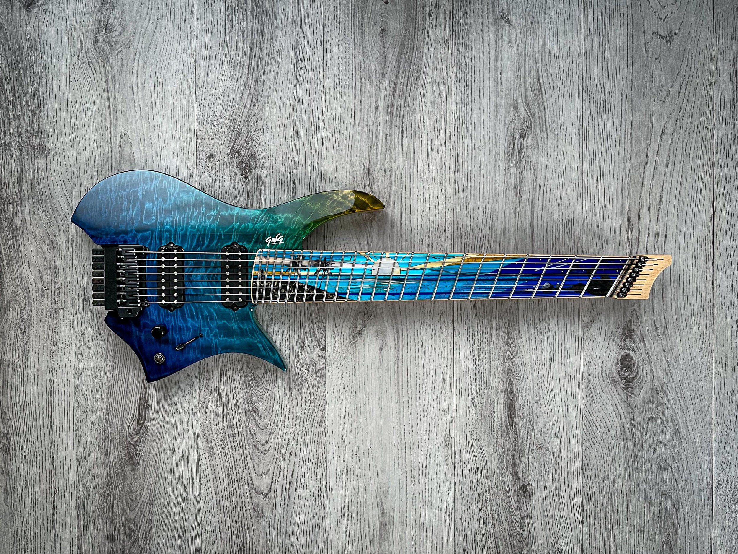 GNG Giulio Negrini electric guitar with a blue-green body, black pickups, and a multicolored fretboard, lying on a gray wooden floor.