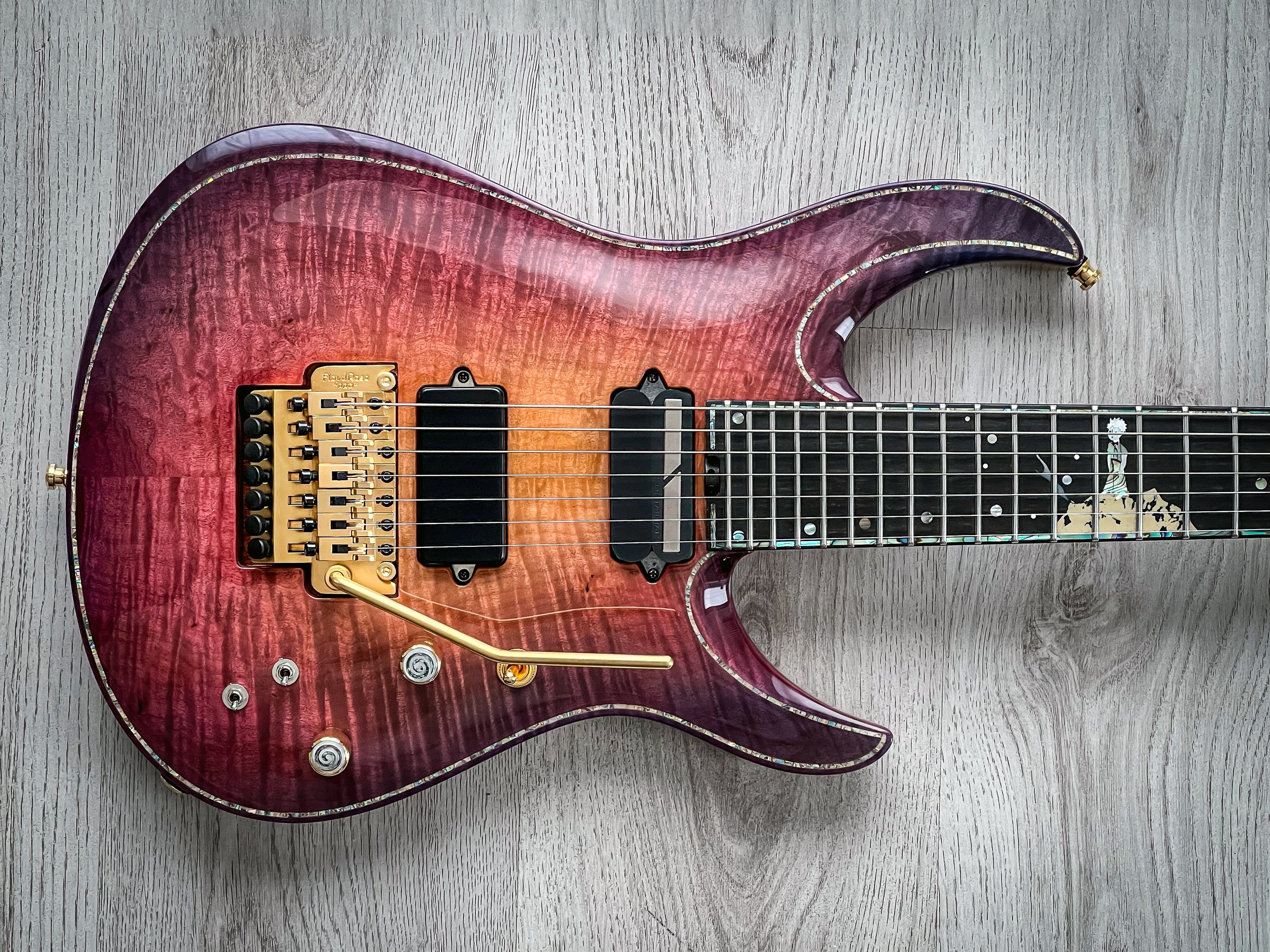 A pink to orange gradient GNG Giulio Negrini electric guitar with a gold Floyd Rose tremolo system and black pickups, lying on a light wood surface.