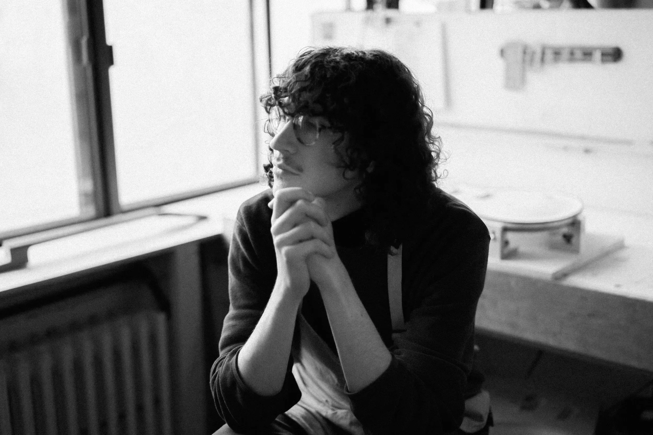 William Parent Senez, a young man with curly hair and glasses, is sitting thoughtfully by a window in a room with a table and a guitar case in the background.