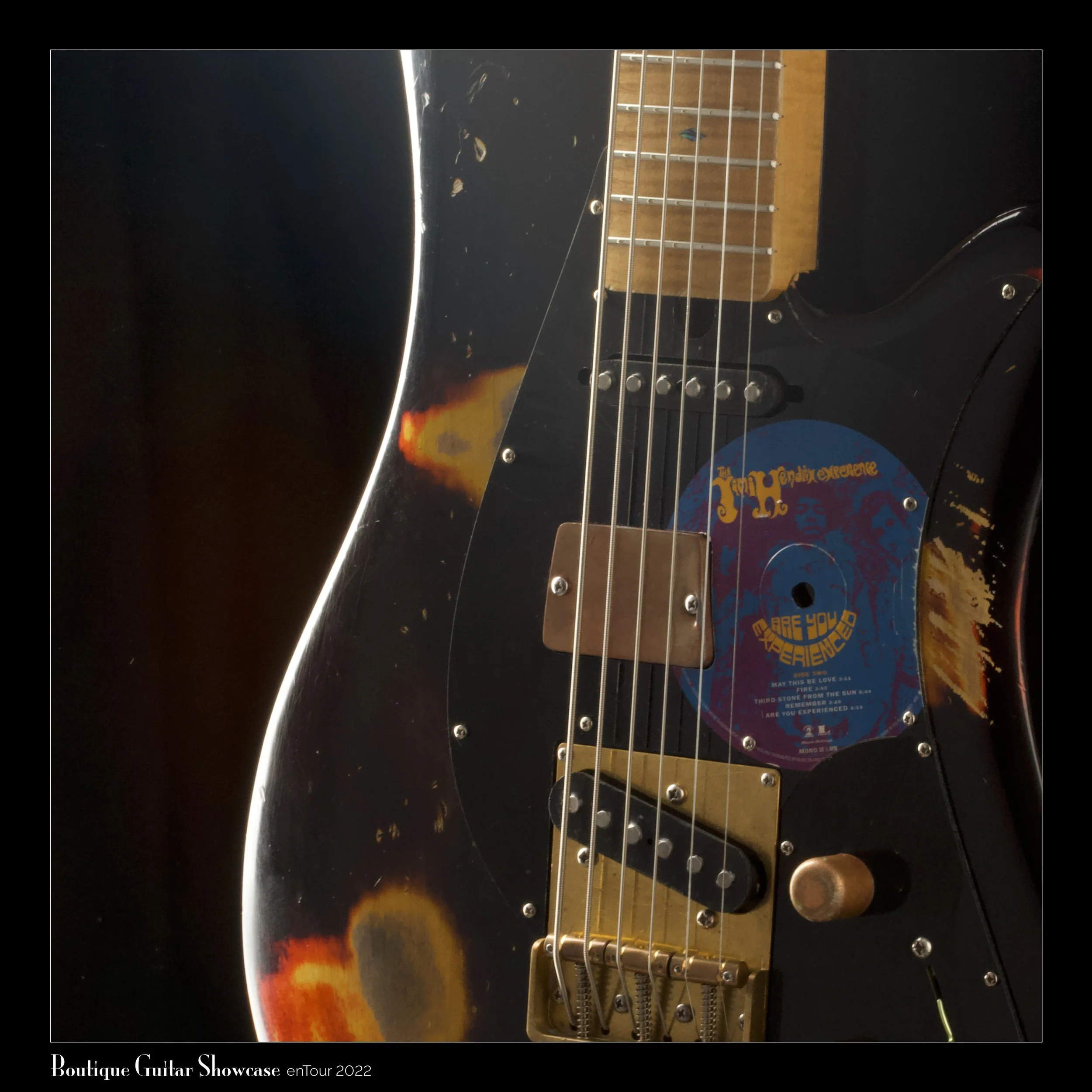 Close-up of a Bacci Baritone electric guitar with a worn, distressed finish, featuring a sticker resembling the cover of Jimi Hendrix's album 'Are You Experienced?' on the body. Part of the Boutique Guitar Showcase event from enTour 2022.