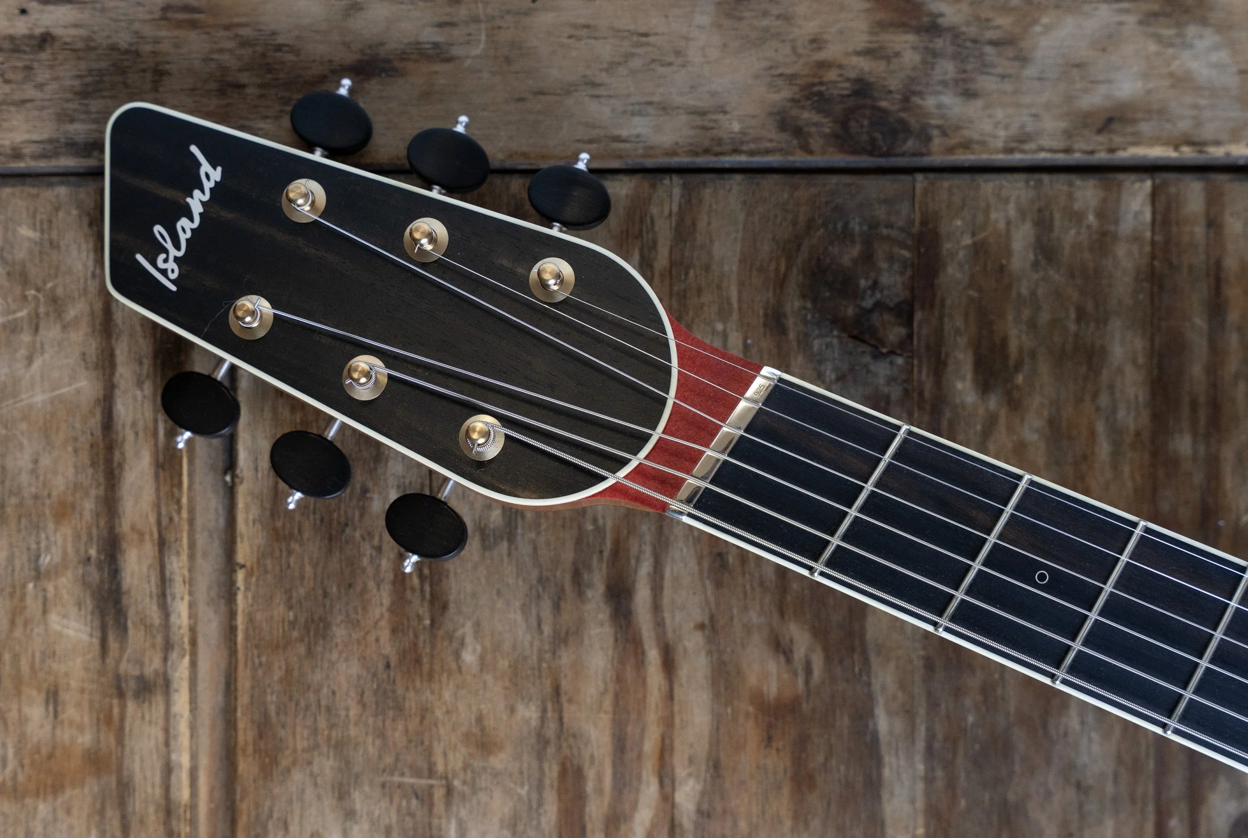 Close-up of the headstock of an Island Instrument brand acoustic guitar, showing tuning pegs, strings, and part of the fretboard, placed on a wooden surface.
