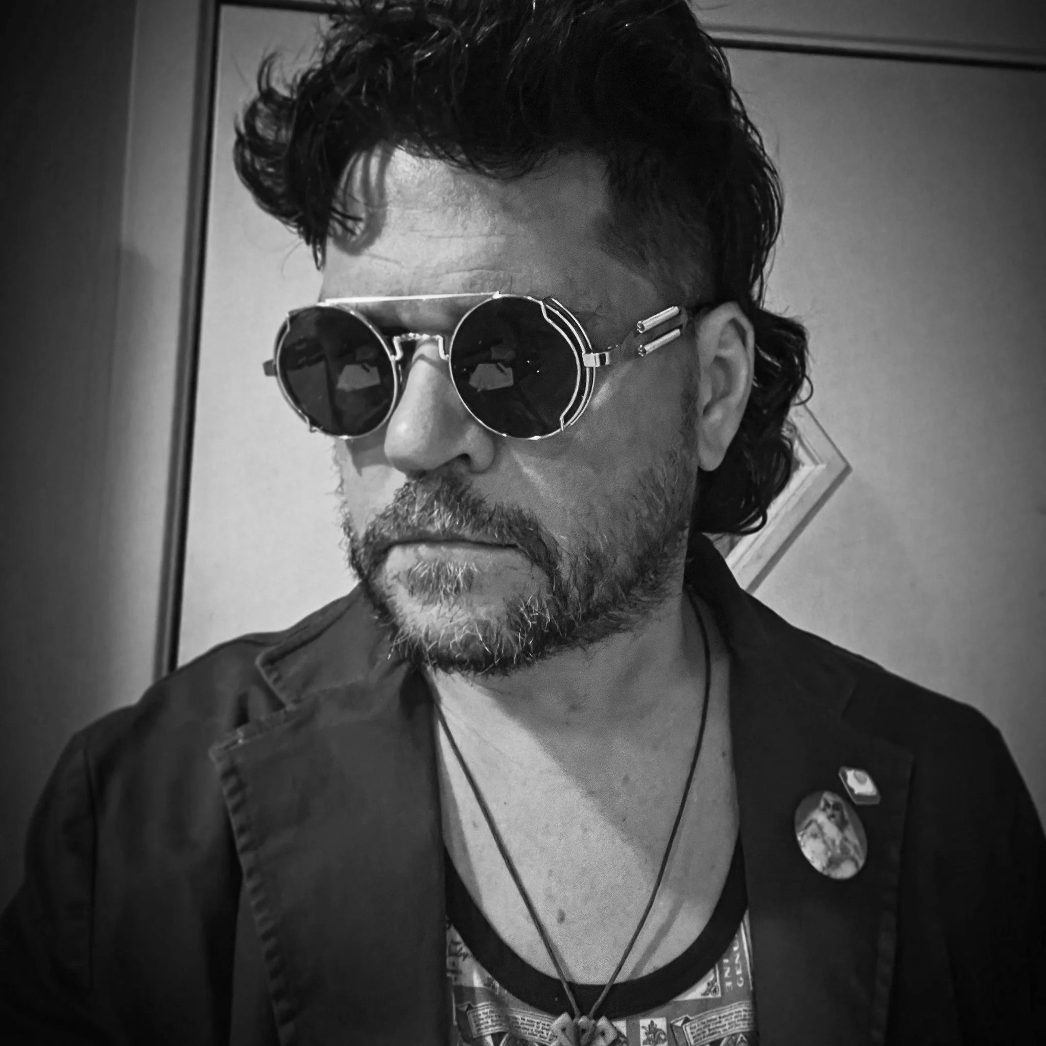 Black and white photo of Jeremy Jenkins wearing dark sunglasses, with wavy hair, a beard, and a serious expression. He has various pins and accessories on his clothing and is looking slightly downward.