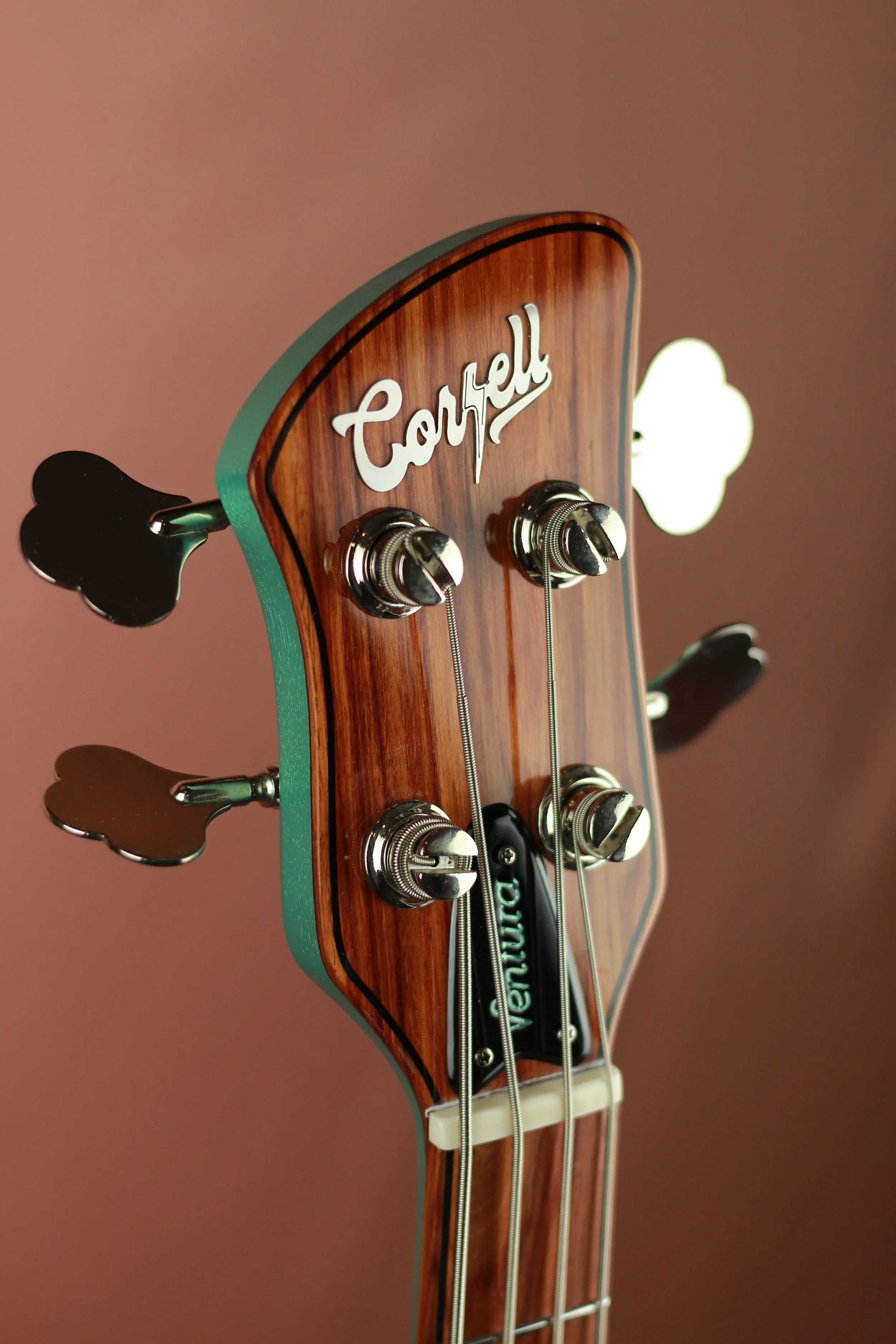 Close-up of a Corsell Electric bass headstock with tuning pegs, strings, and a strap block, against a blurred pink background.
