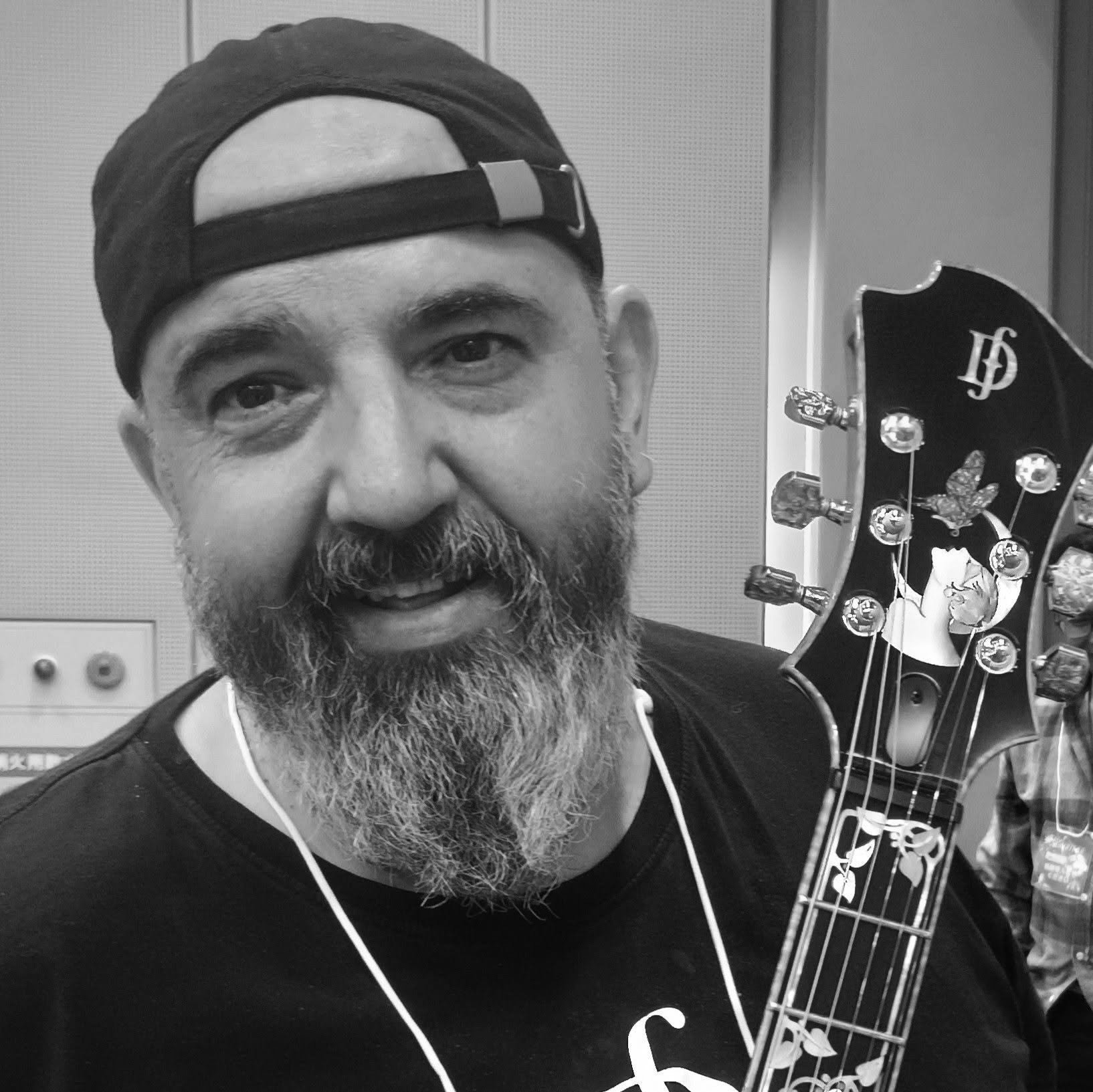 Black and white photo of Emil Djerjinski with a beard wearing a backwards cap holding a guitar headstock with tuning pegs.