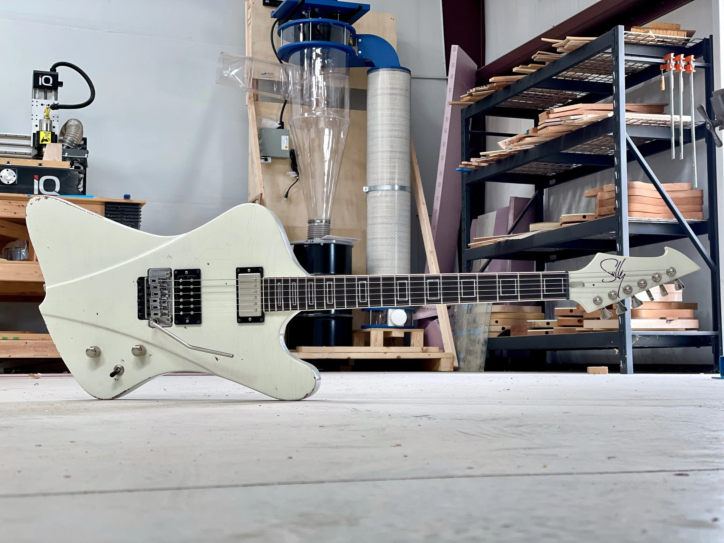 White electric Sully guitar lying on its side on a wooden workshop floor with woodworking tools and shelves in the background.