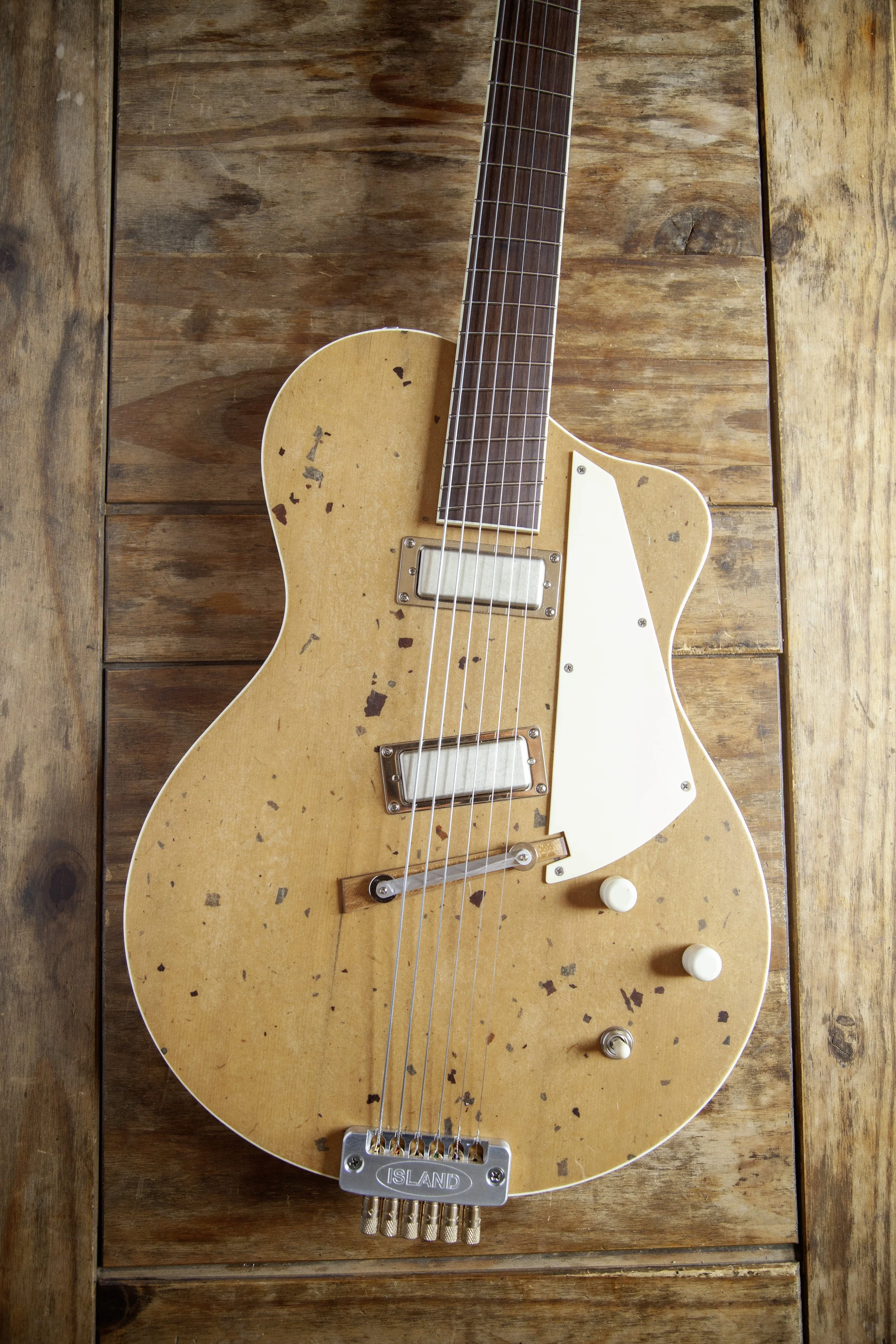 A vintage, worn electric guitar with a natural wood finish, resting on a wooden surface. The guitar has a white pickguard, two pickups, and control knobs. It features a bridge with multiple gold-colored strings and a metal bridge assembly  "ISLAND." 