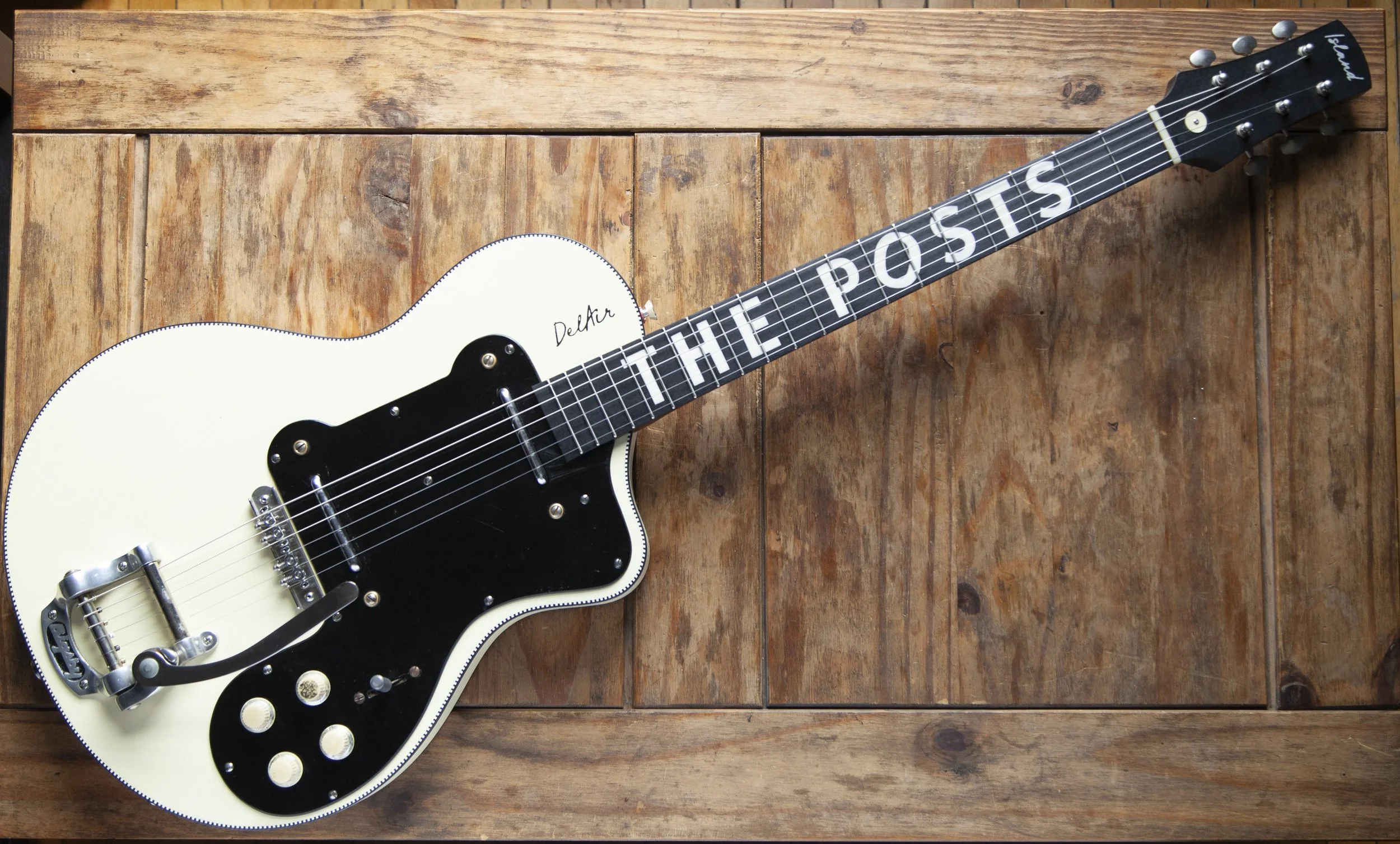 A white electric Island Instruments guitar with black pickguard and the words 'THE POSTS' on the fretboard, resting on a wooden surface.