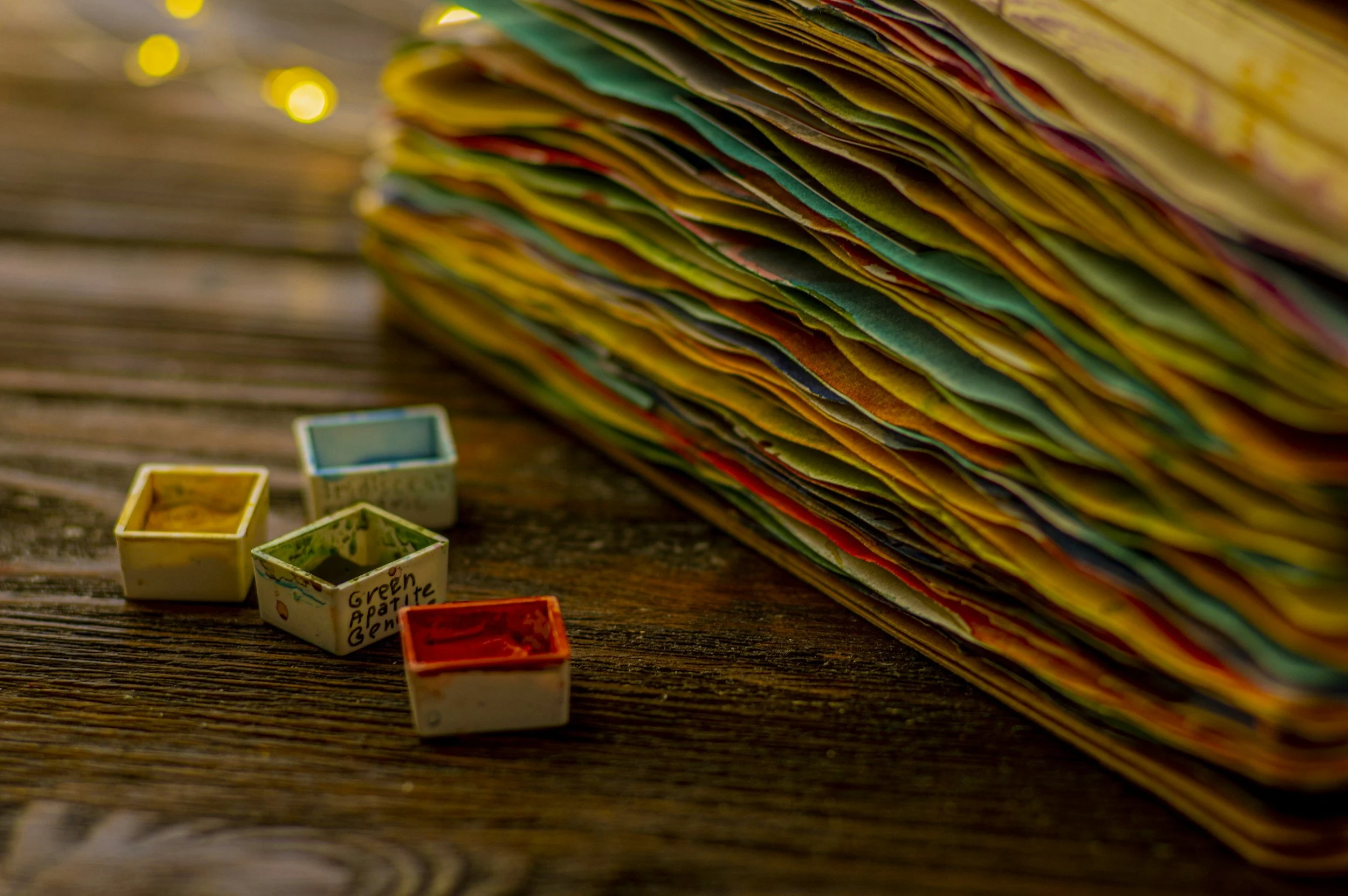 A large stack of colorful paper or fabric scraps on a wooden surface, with four small square paint containers in front, some with paint inside and some labeled, like "Green Apple".