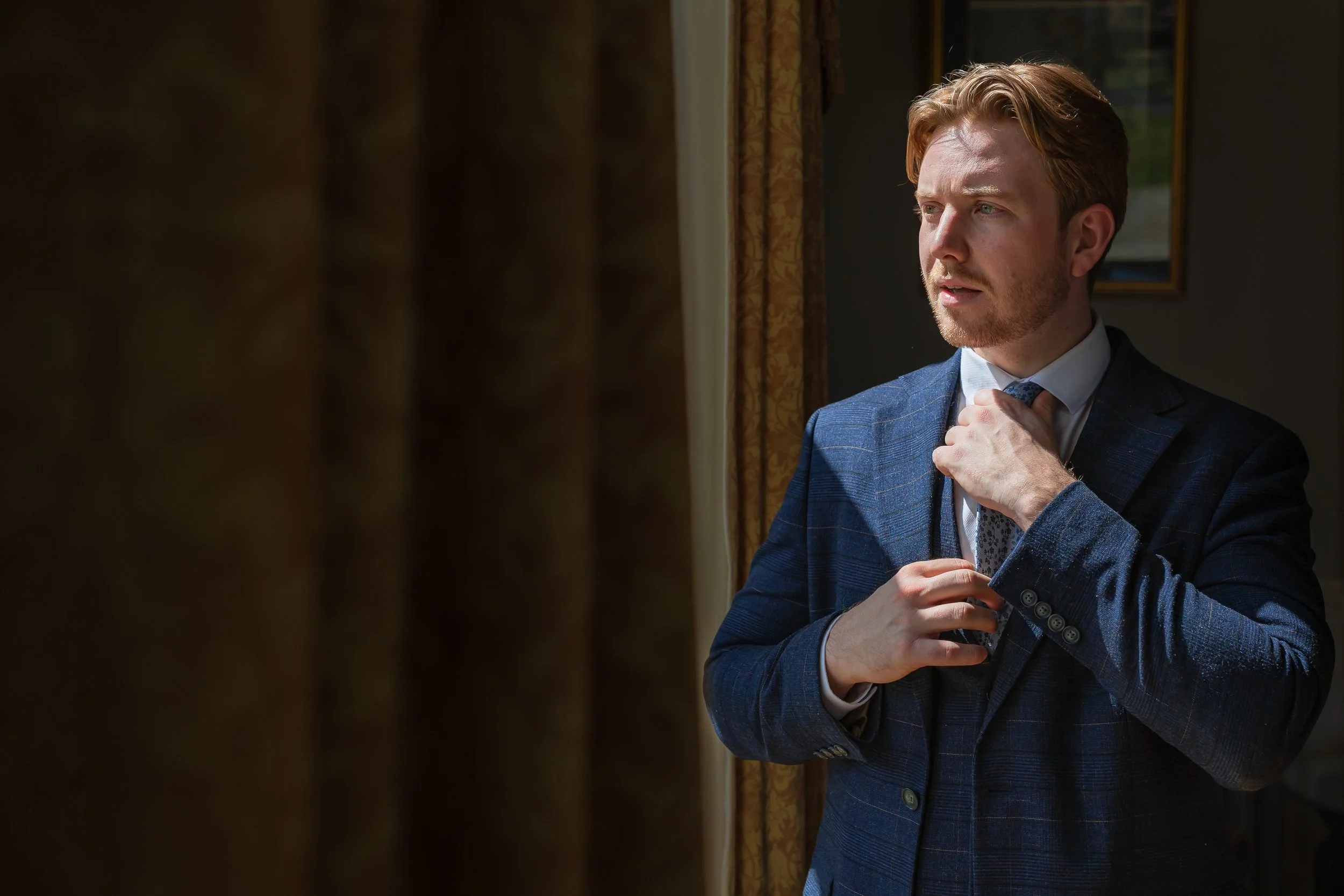 Groom adjusting his tie in window light during morning preparations at Eaves Hall in Lancashire