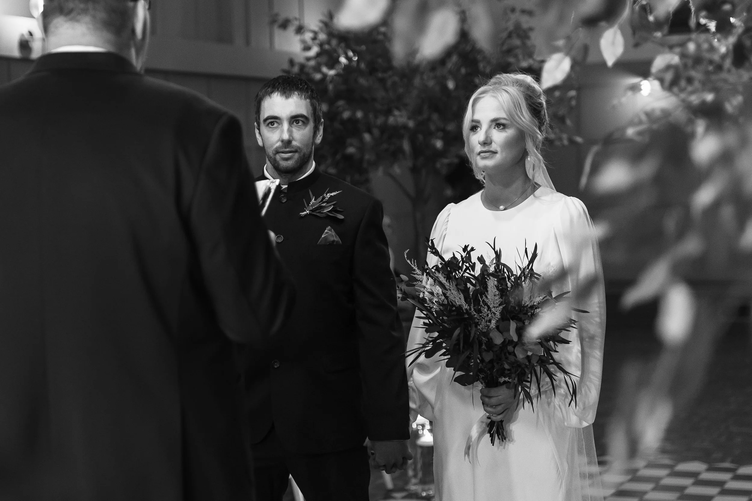 Black and white wedding ceremony at Larkspur Lodge with bride and groom holding hands during vows, framed through soft foliage.