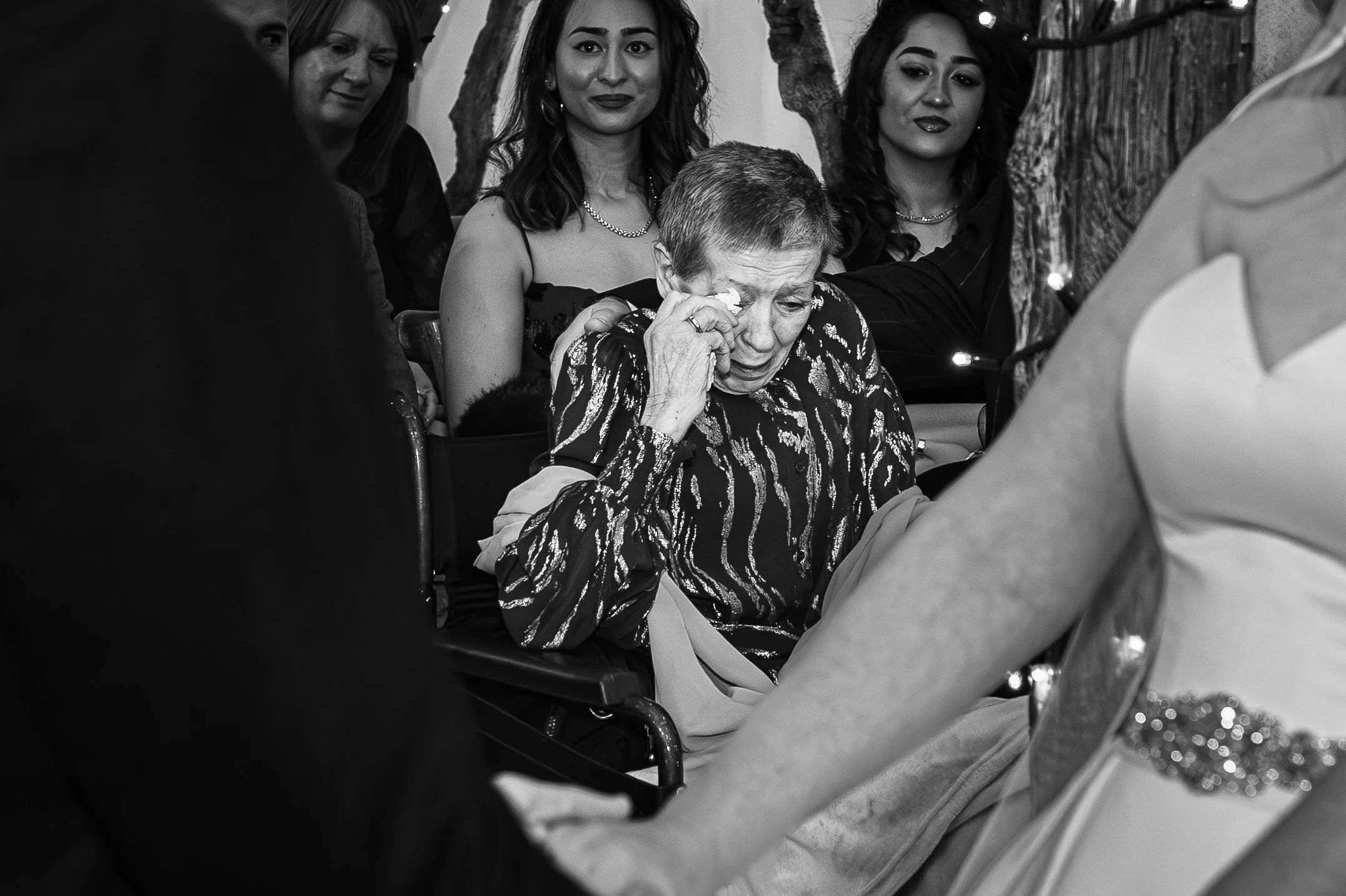 Black and white documentary wedding ceremony moment showing a grandmother in tears while the bride and groom hold hands in the foreground.