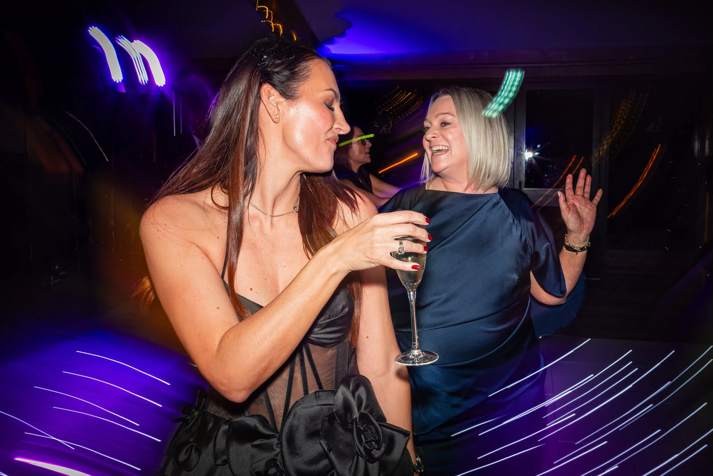 Two women dancing and laughing at a party with colourful motion-blur lights, captured candidly by a Cheshire event photographer.