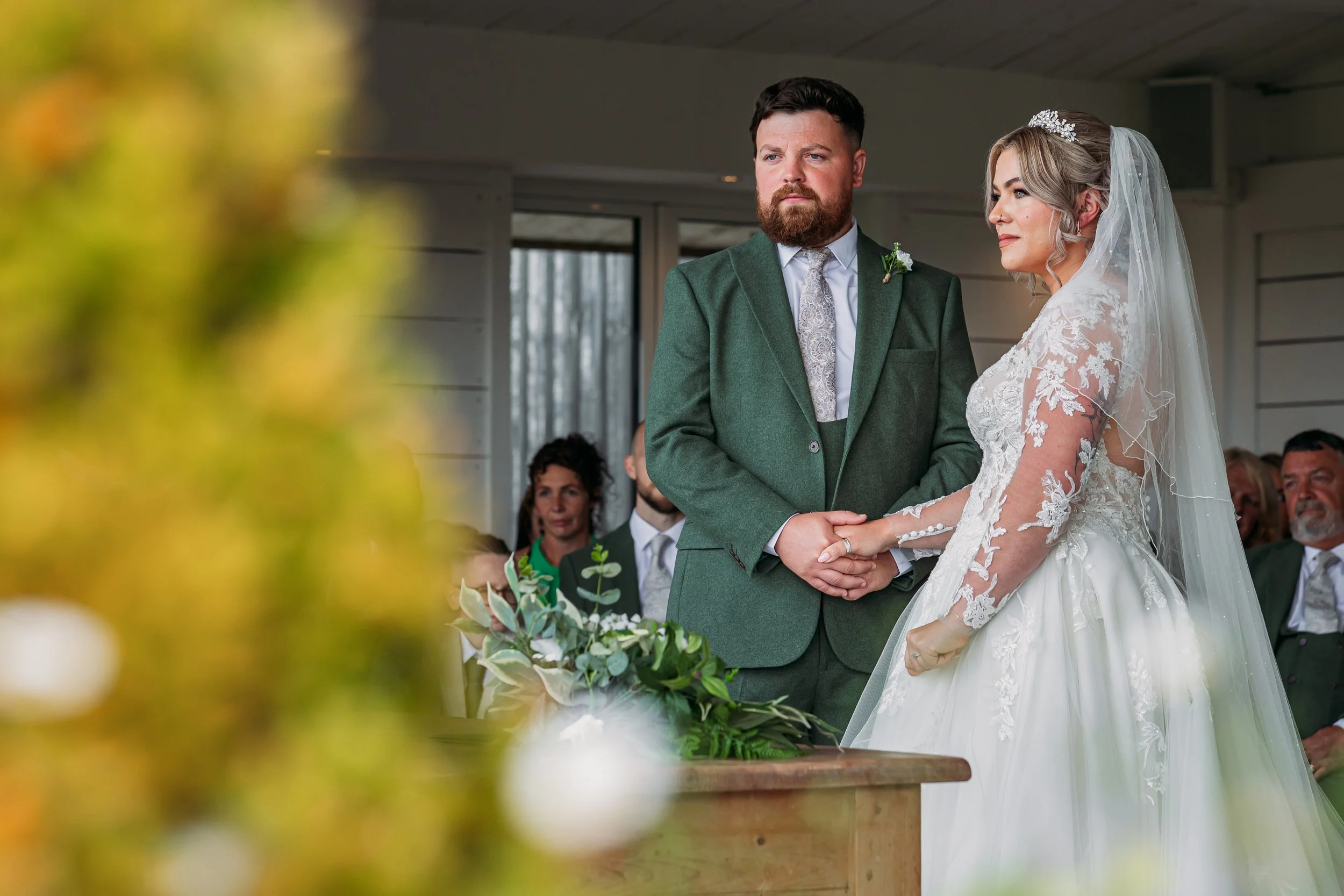 Bride and groom holding hands during wedding ceremony at Bashall Barn with soft floral foreground and natural light.