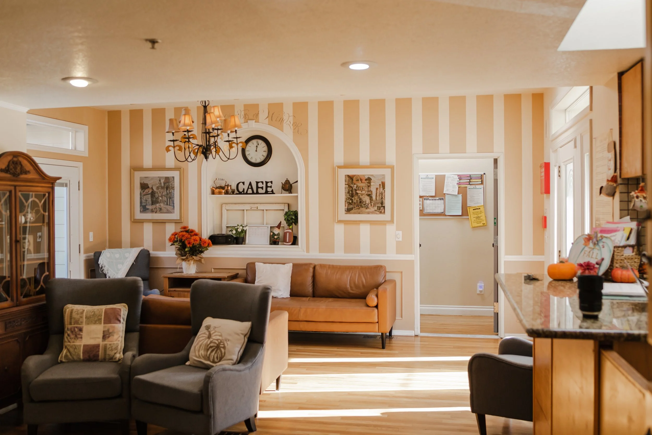Cozy living room with striped walls and mixed furniture, including a leather sofa, patterned armchairs, and a chandelier. Decorative wall features include artwork, a clock, and a shelf labeled 'Cafe.' Wooden accents, flowers, and seasonal decor add warmth to the space.