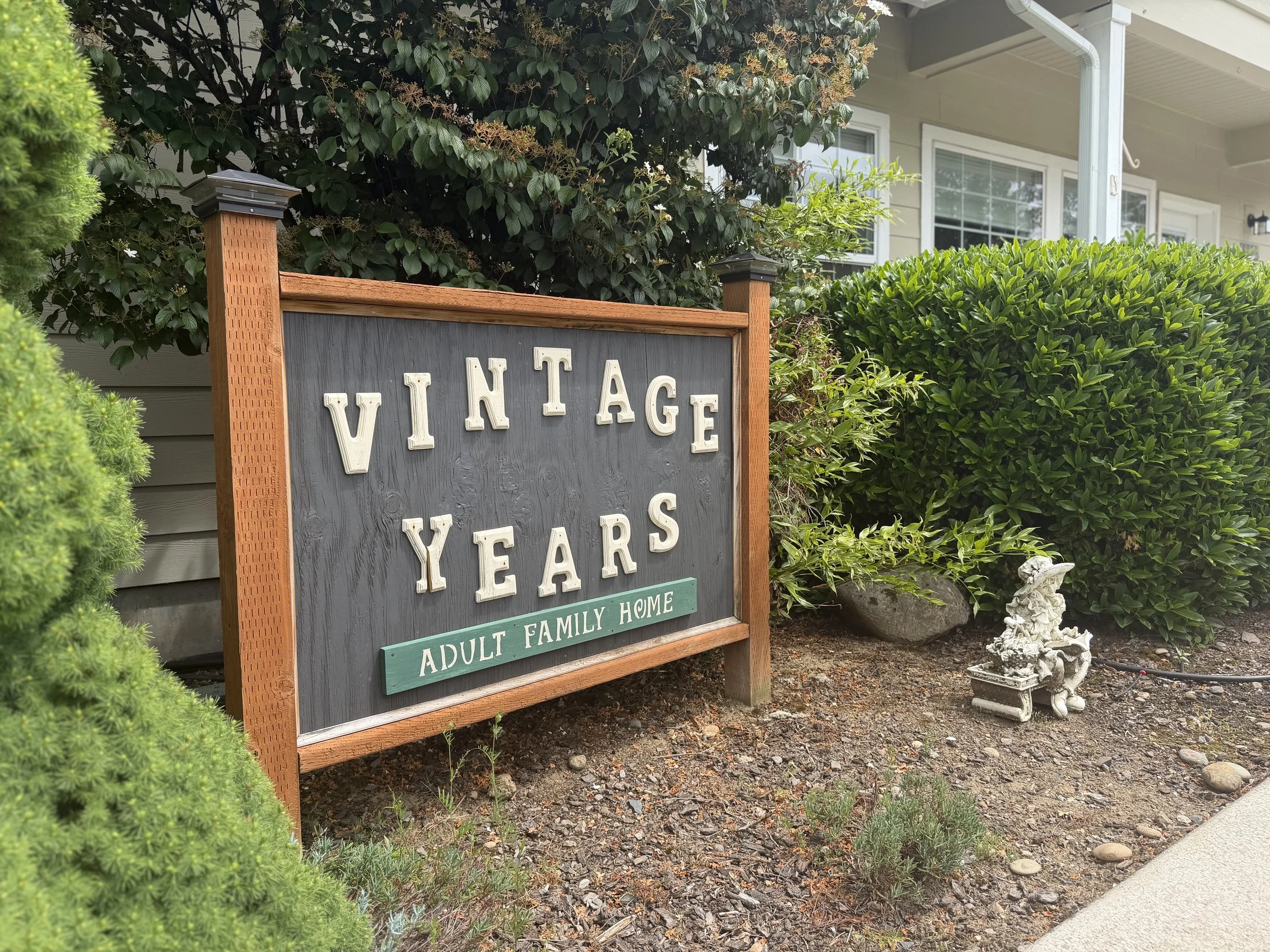 Sign reading "Vintage Years Adult Family Home" in front of lush greenery and a building with windows.