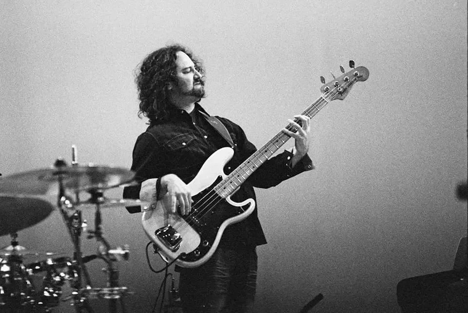 A man with curly hair and a beard playing a bass guitar in a music studio, in black and white.