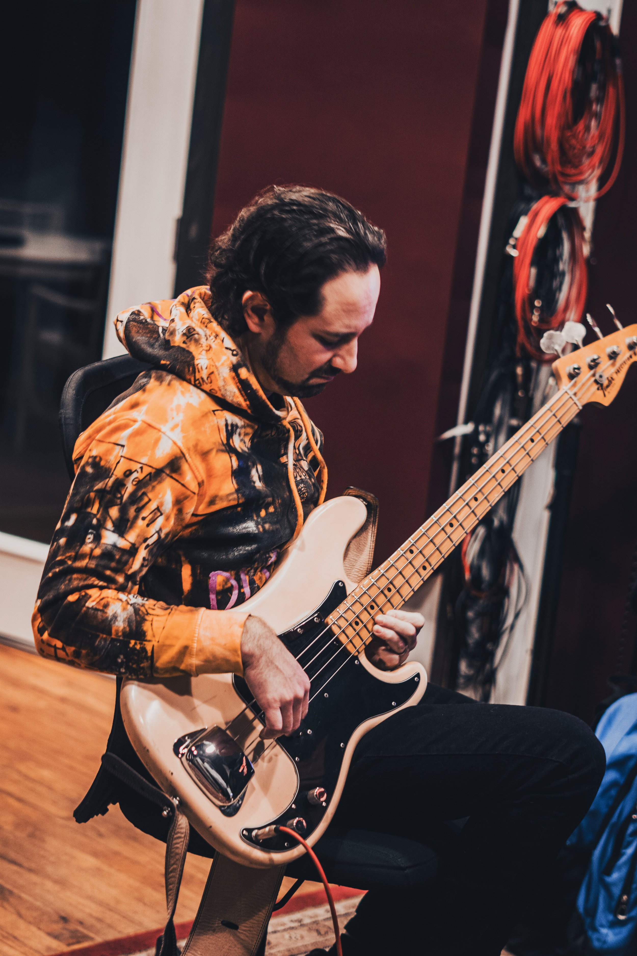 A man with dark hair, wearing a orange and black hoodie, playing an electric bass guitar in a recording studio.