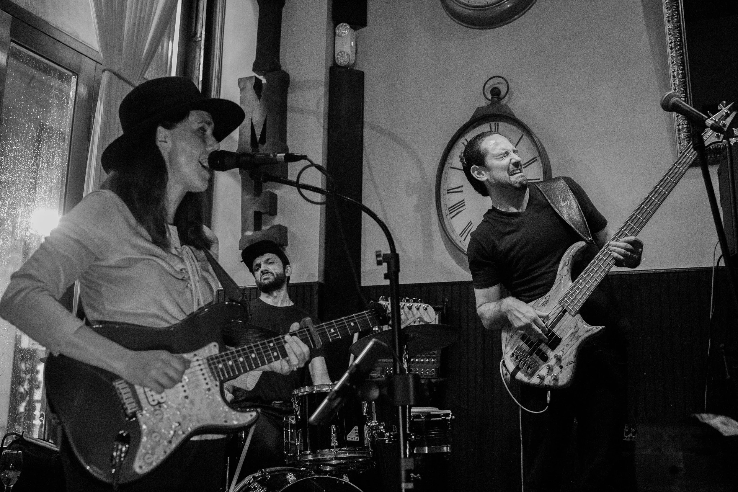 A black-and-white photo of three musicians performing in an indoor venue. A woman on the left plays an electric guitar and sings into a microphone, wearing a hat. A man on the right plays a bass guitar with a pained expression. A man in the backgroun