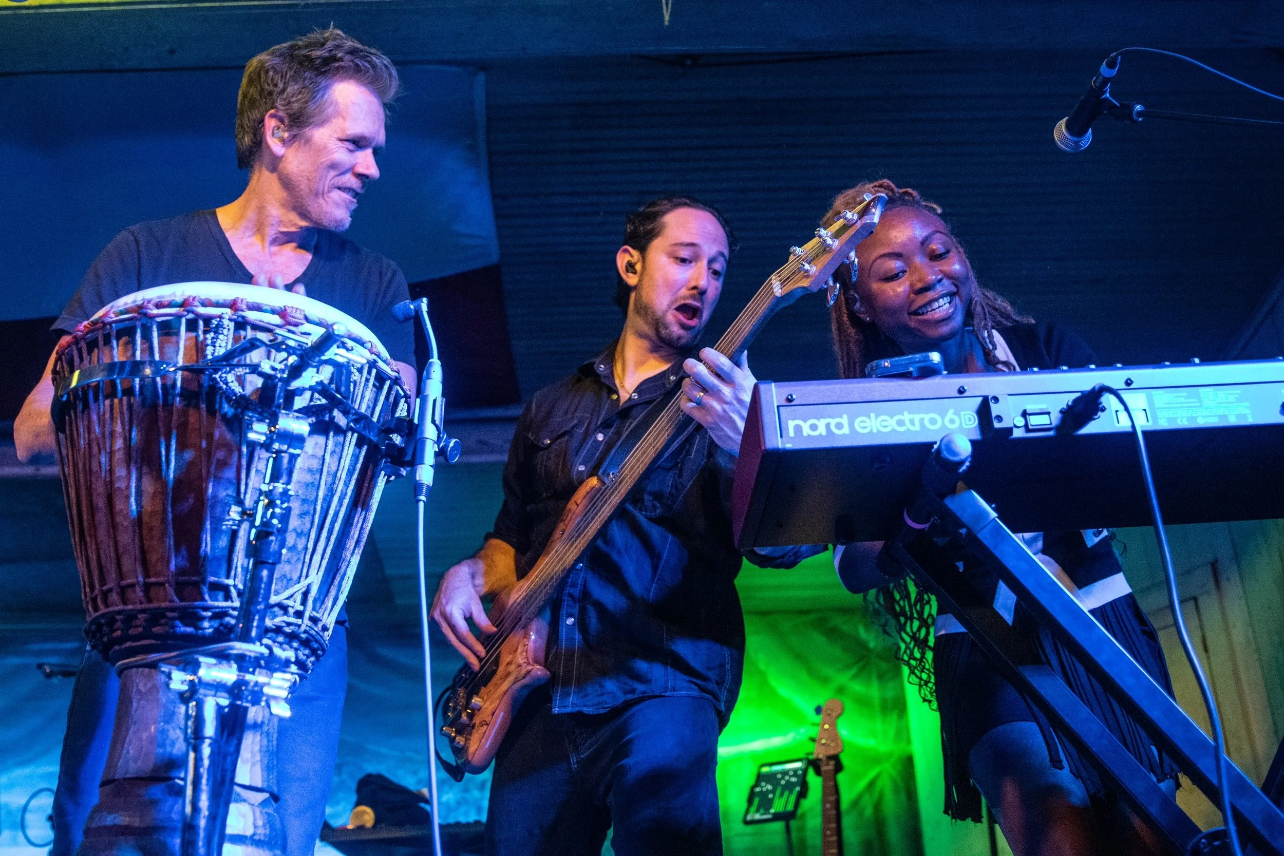 Three musicians performing together on stage: a man playing a hand drum, a man playing an electric bass, and a woman playing a keyboard.