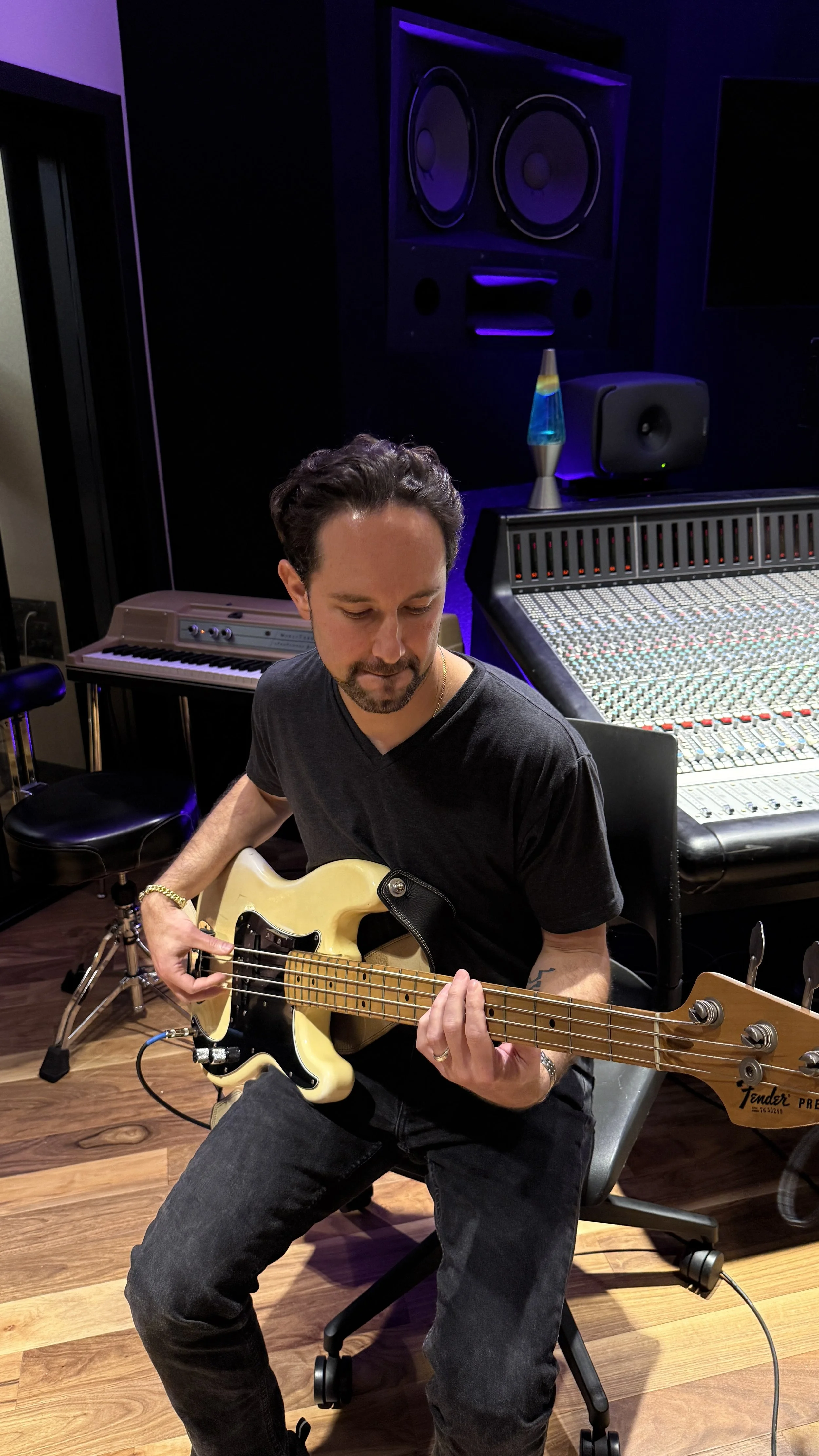 A man in a black T-shirt playing a yellow electric bass guitar in a music studio with a mixing console, speaker, and lava lamp in the background.