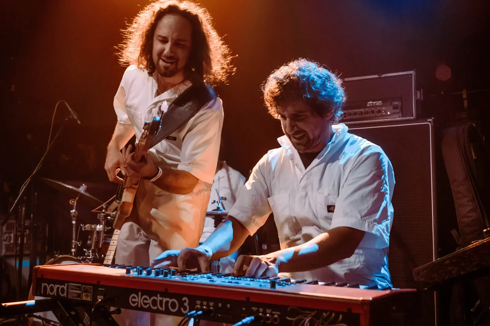 Two musicians performing on stage, one playing a guitar and the other using a keyboard, both wearing white outfits, with warm stage lighting.