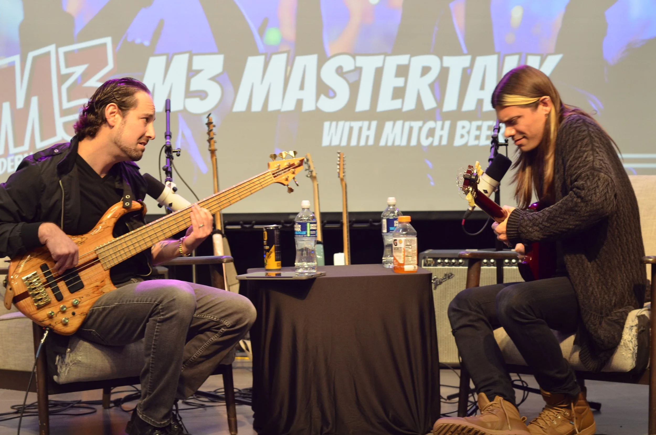 Two musicians sit on stage with guitars, performing in front of a screen that reads 'M3 Masterclass with Mitch Beer.' The man on the left plays a bass guitar, and the man on the right plays an acoustic guitar. They have water bottles and coffee on a 