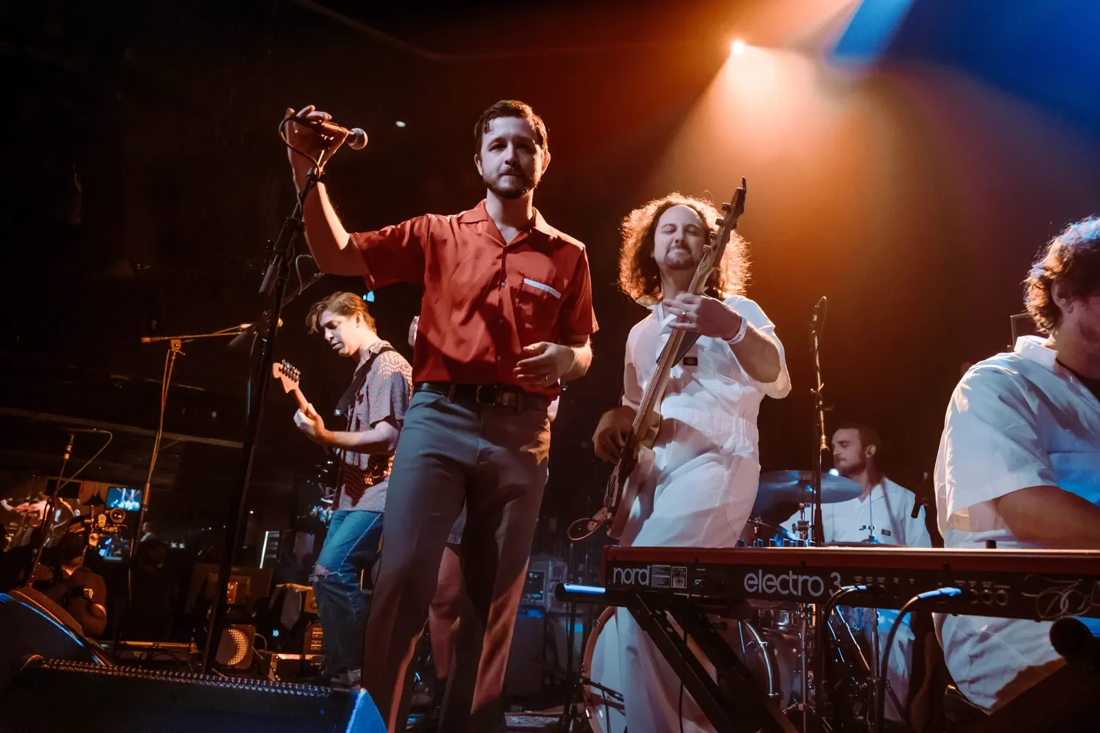 Band performing live on stage, featuring a vocalist holding a microphone, a guitarist, a bassist, a keyboardist, and a drummer, with stage lighting creating an energetic atmosphere.