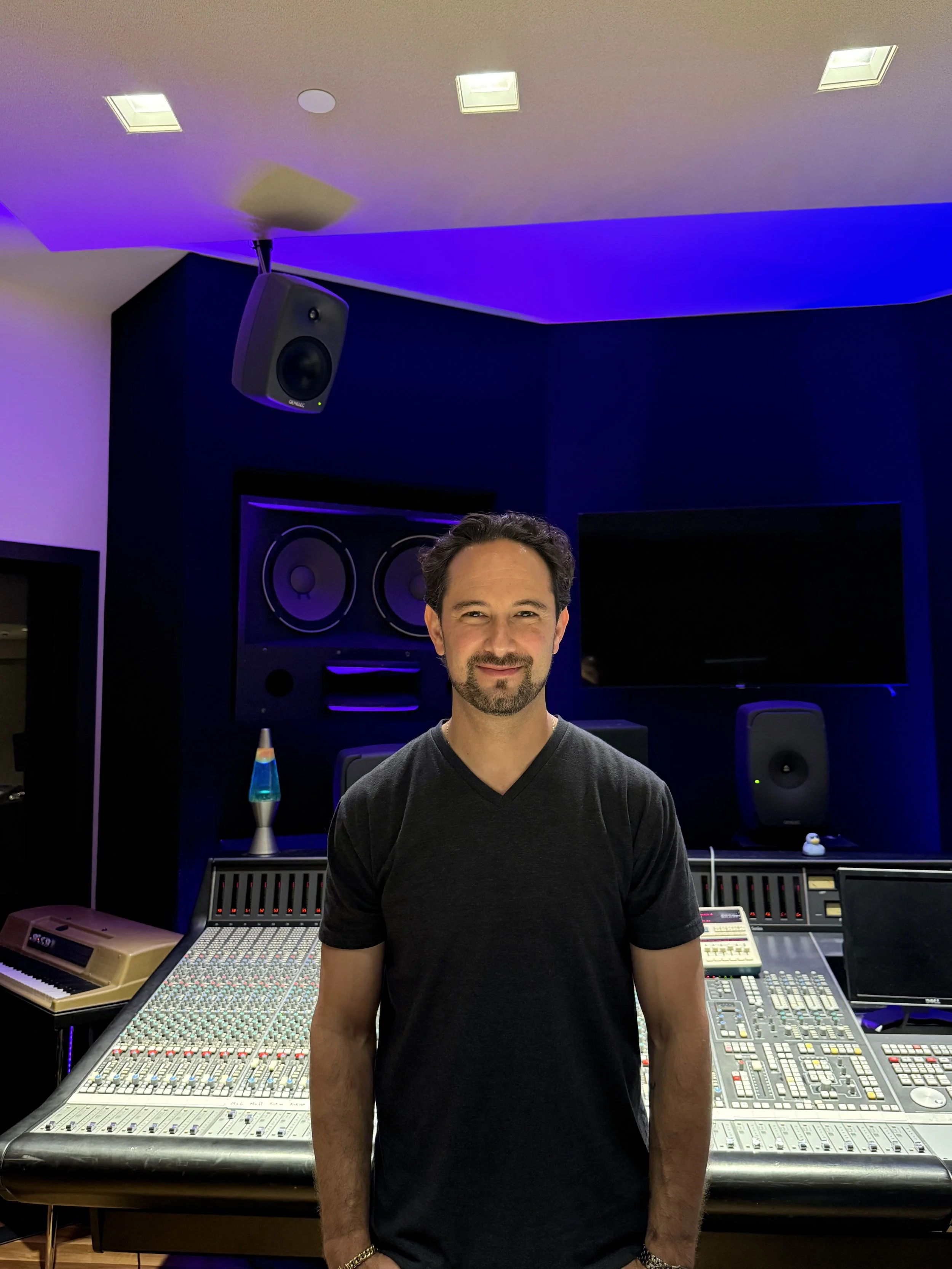 A man with dark hair and a beard standing in a music recording studio, surrounded by sound mixing equipment, with blue and purple lighting.