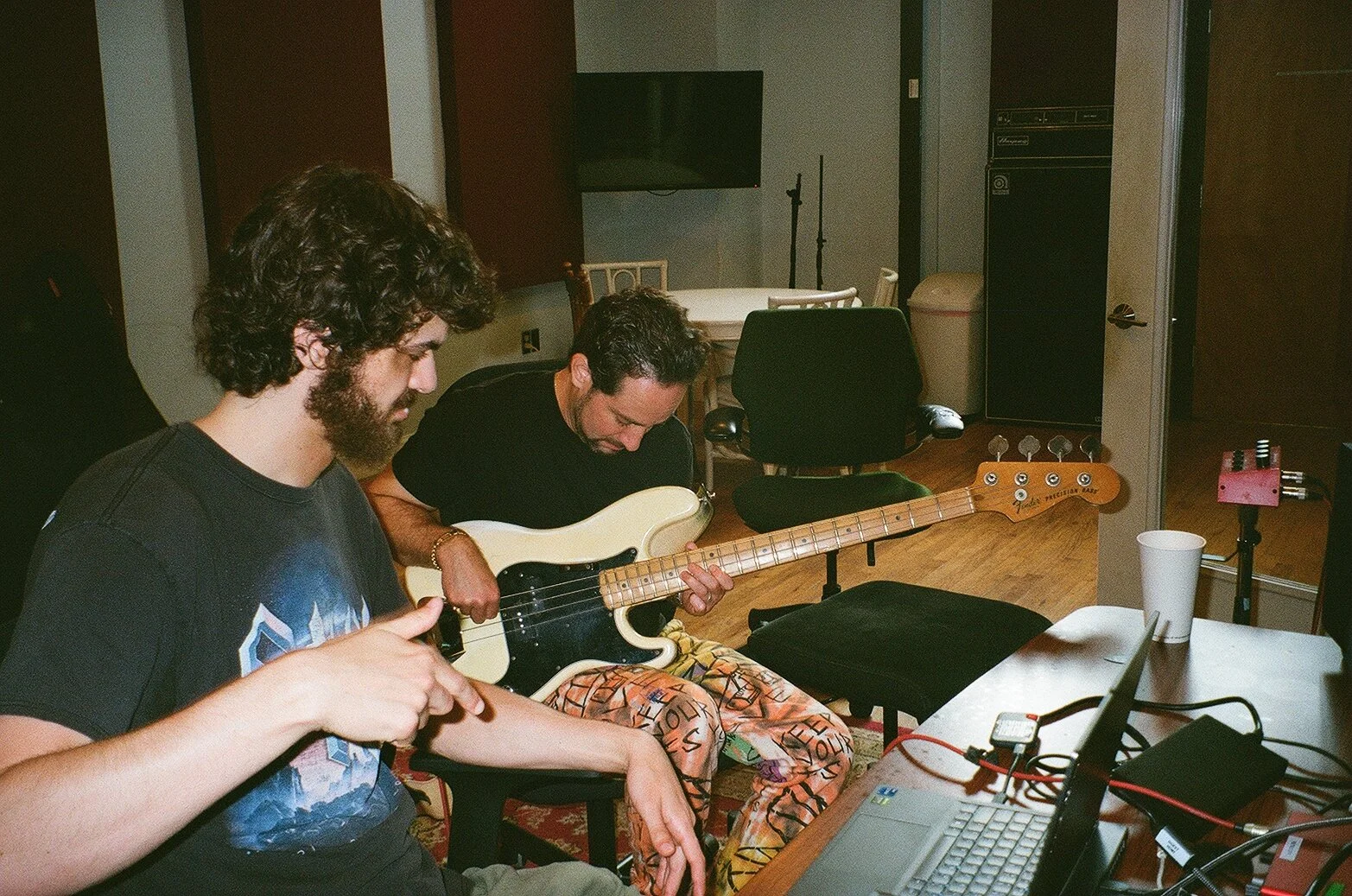 Two men in a music studio, one with curly hair and a beard wearing a black t-shirt, the other with short hair and a beard wearing colorful pants, sitting at a table with music equipment and a guitar.