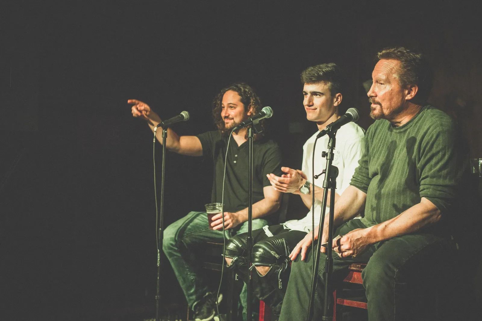 Four men sitting on a stage with microphones, two of them are clapping, one pointing, and one has a drink, in a dark setting likely during a live performance or comedy show.