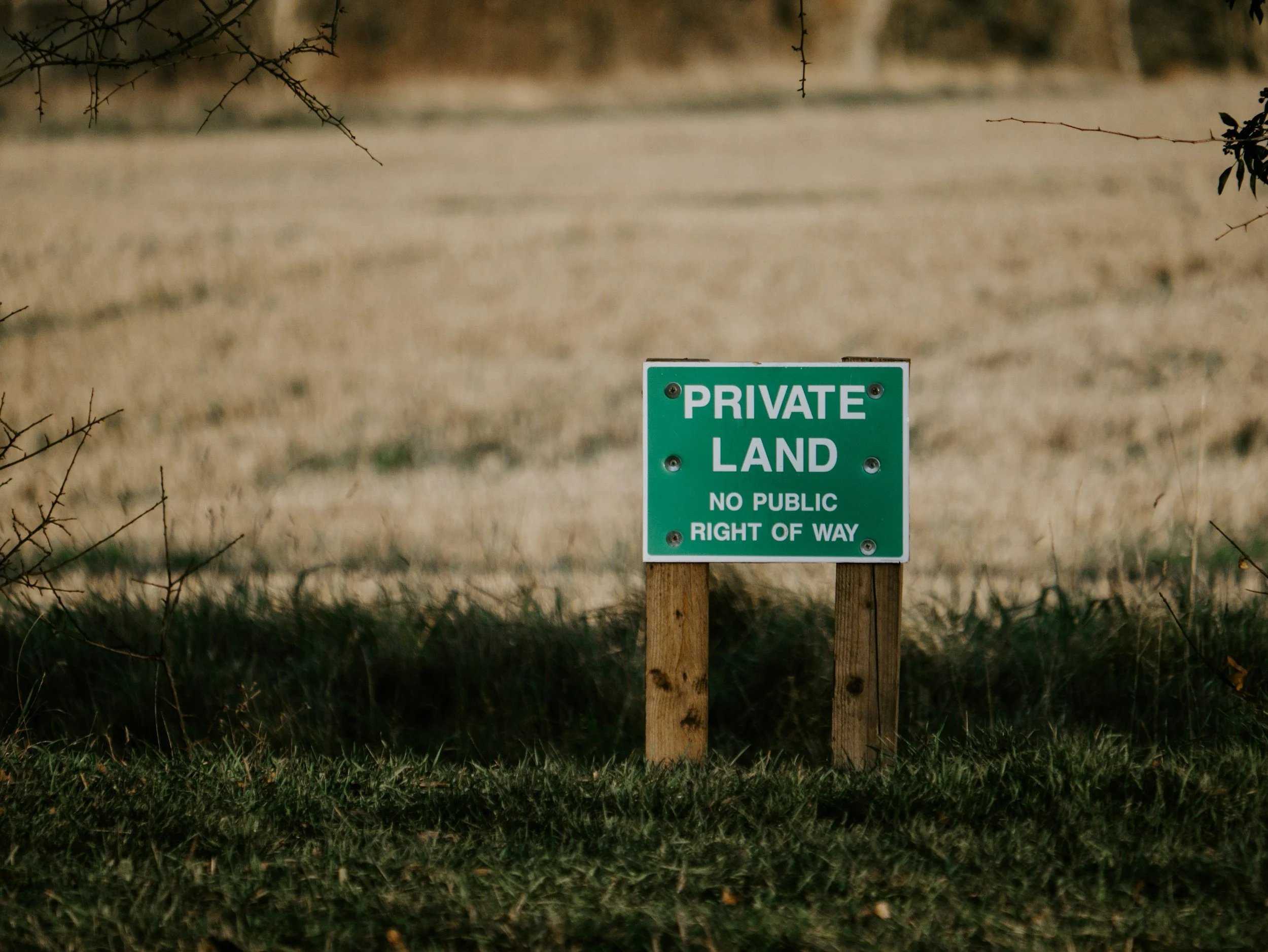 Green sign that says 'PRIVATE LAND NO PUBLIC RIGHT OF WAY' on two wooden posts, in front of a grassy area with a blurred tan field or open space background.
