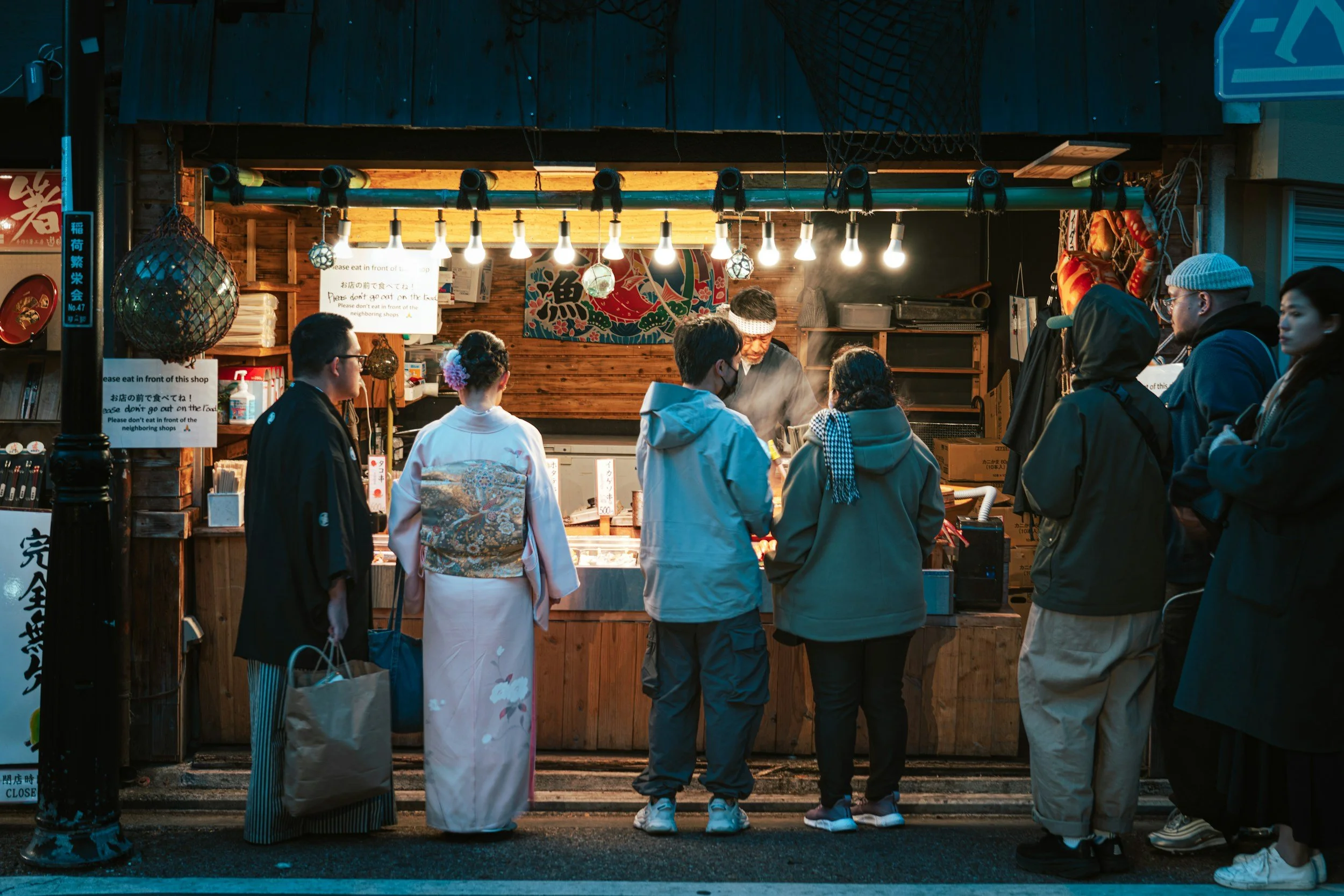 People lined up at a fish stand in a market at night, with bright lights illuminating the scene.
