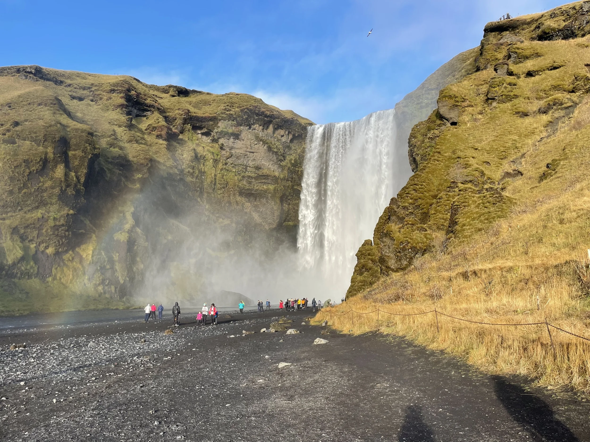 People visiting a waterfall with cliffs covered in moss and grass, a river flowing at the base, and a clear blue sky above.