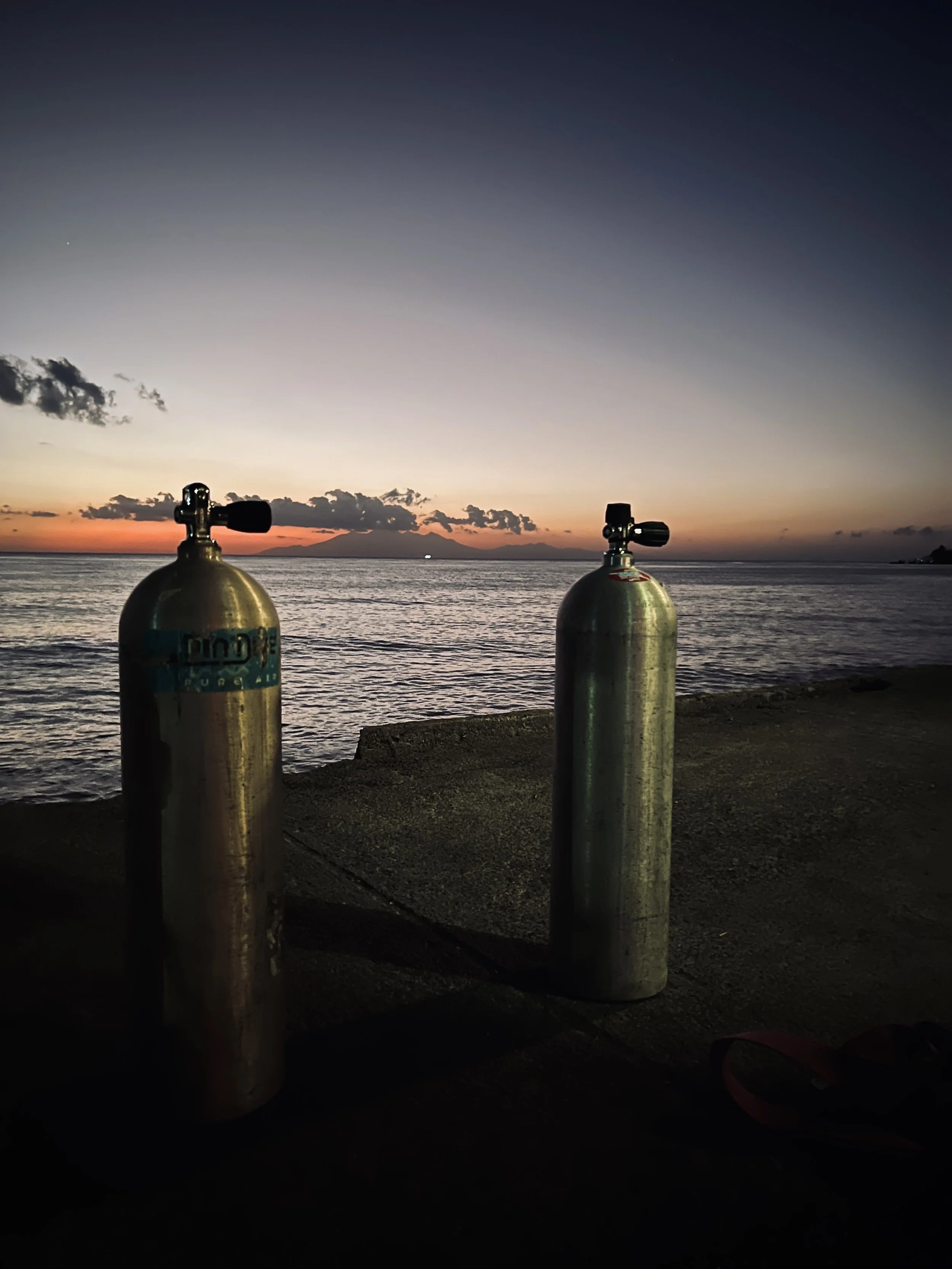 Two scuba tanks on the beach with a view of the ocean at sunset, with clouds and a distant volcanic island in the background.