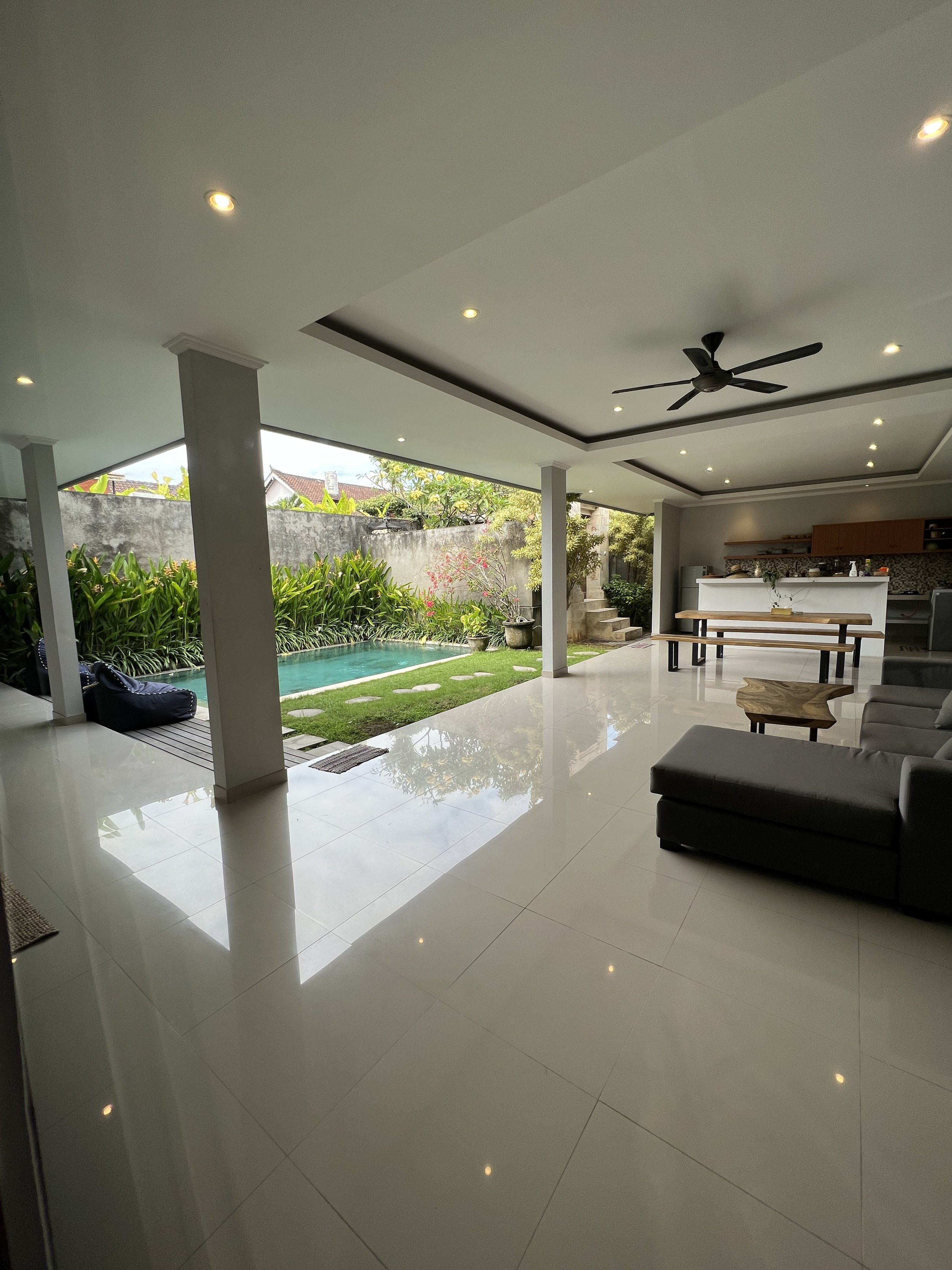 Modern open-plan living space with glossy white tiled floor, white ceiling with recessed lighting, ceiling fan, a view of the garden with a swimming pool, green plants, outdoor furniture, and a kitchen area in the background.