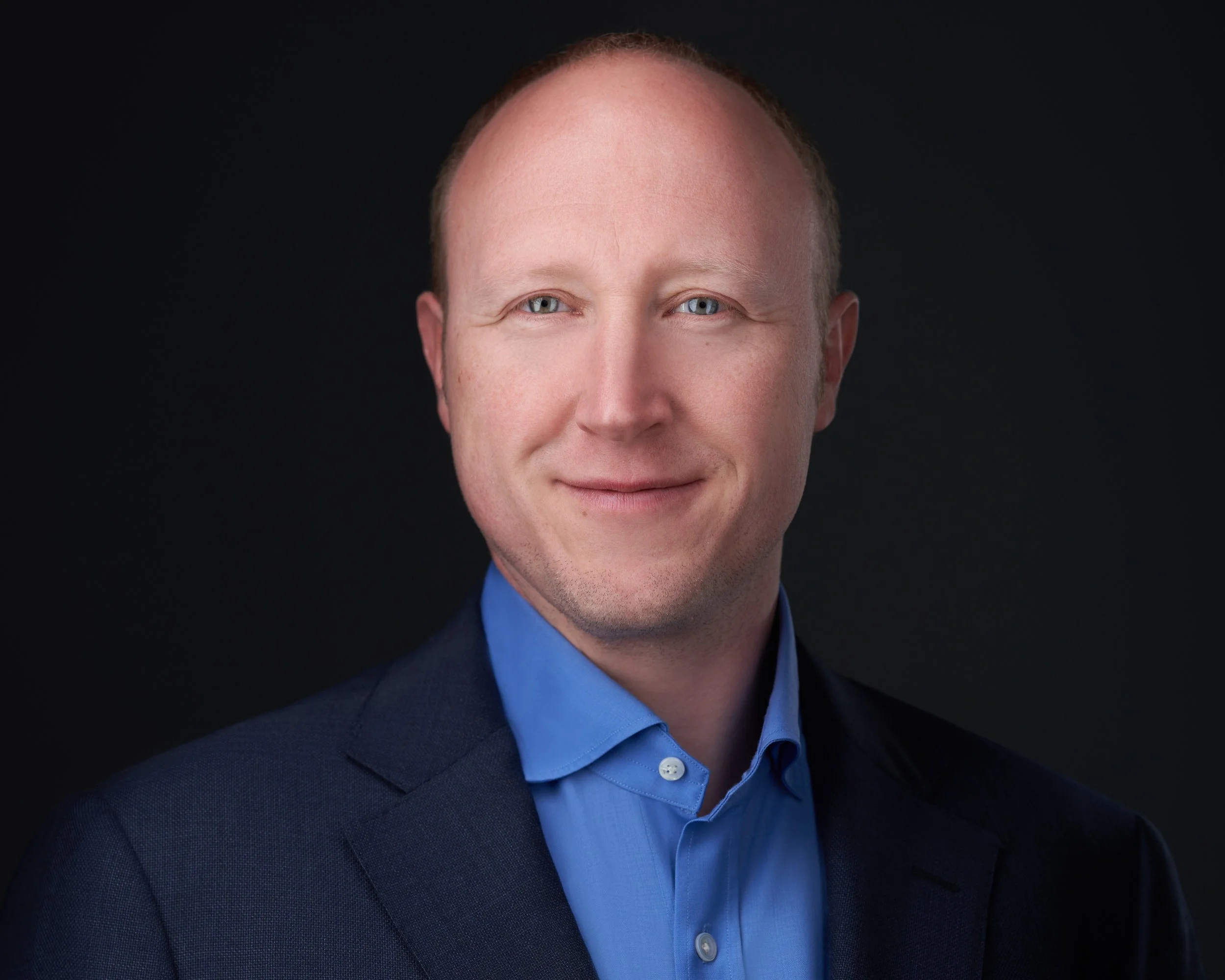 Headshot of a man with light skin, short reddish hair, wearing a dark navy suit and light blue dress shirt, smiling slightly, against a dark background.