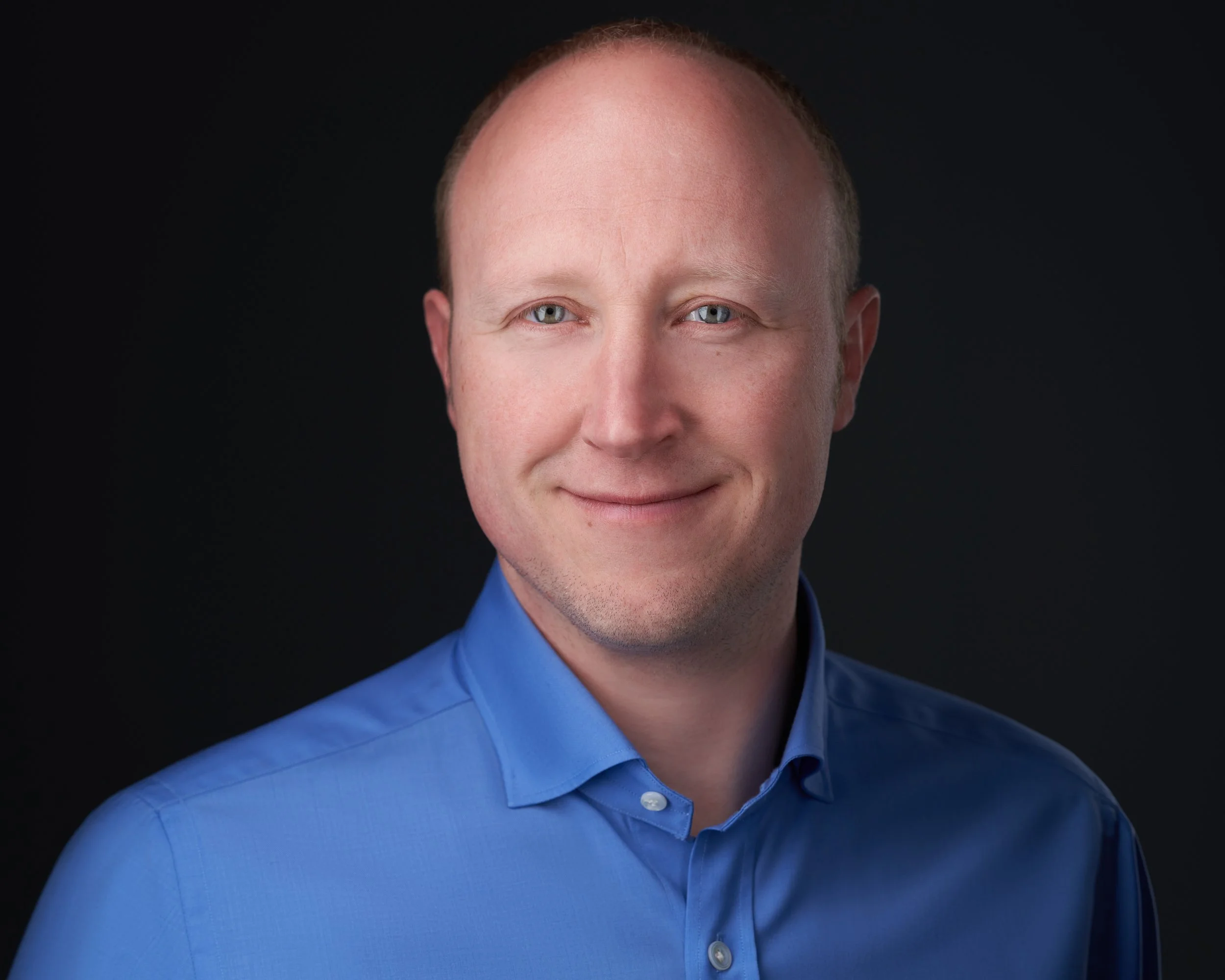 Portrait of a smiling man with short, light brown hair, blue eyes, wearing a blue dress shirt, against a dark background.