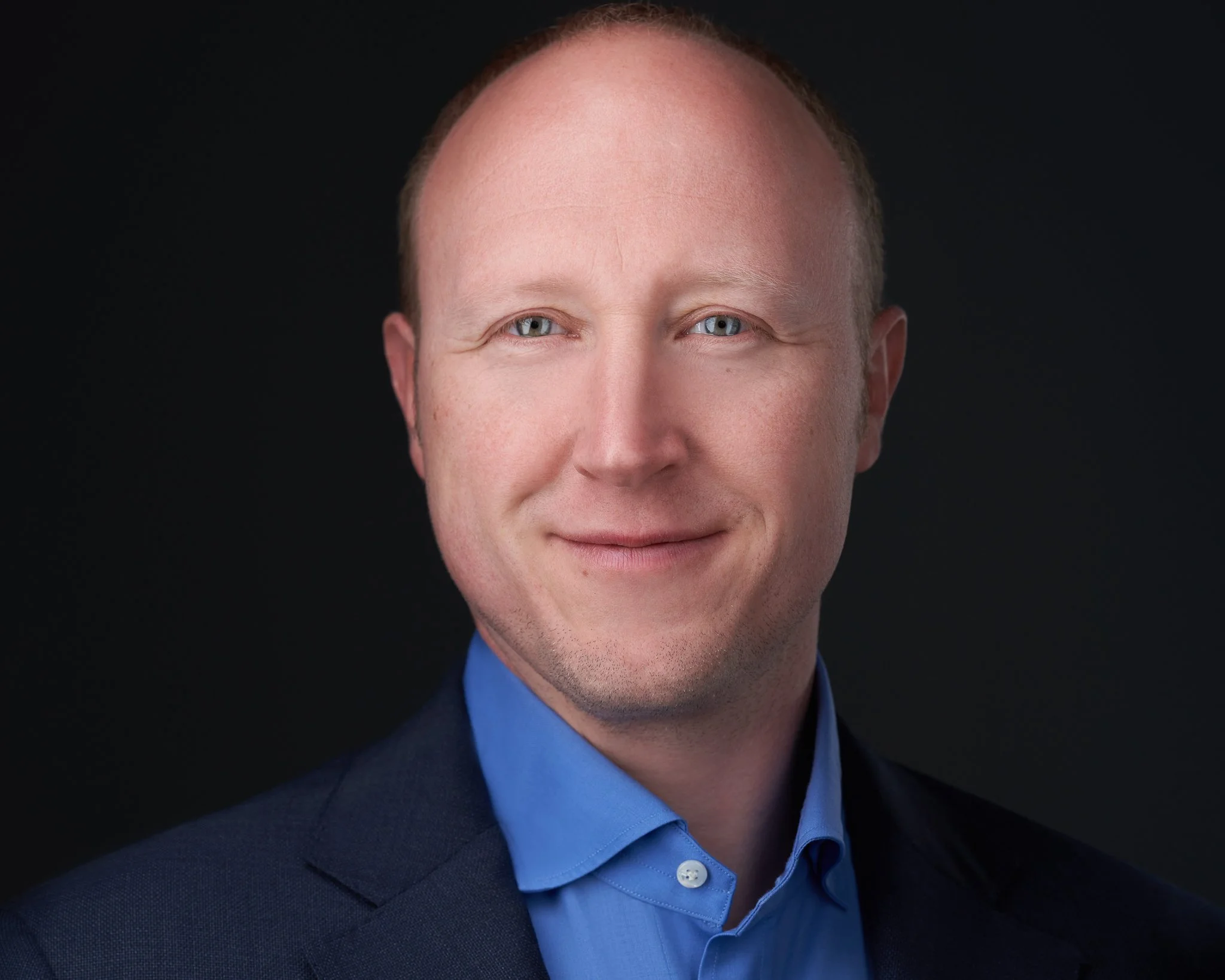Professional headshot of a smiling man with short light brown hair, wearing a dark suit and blue shirt against a black background.