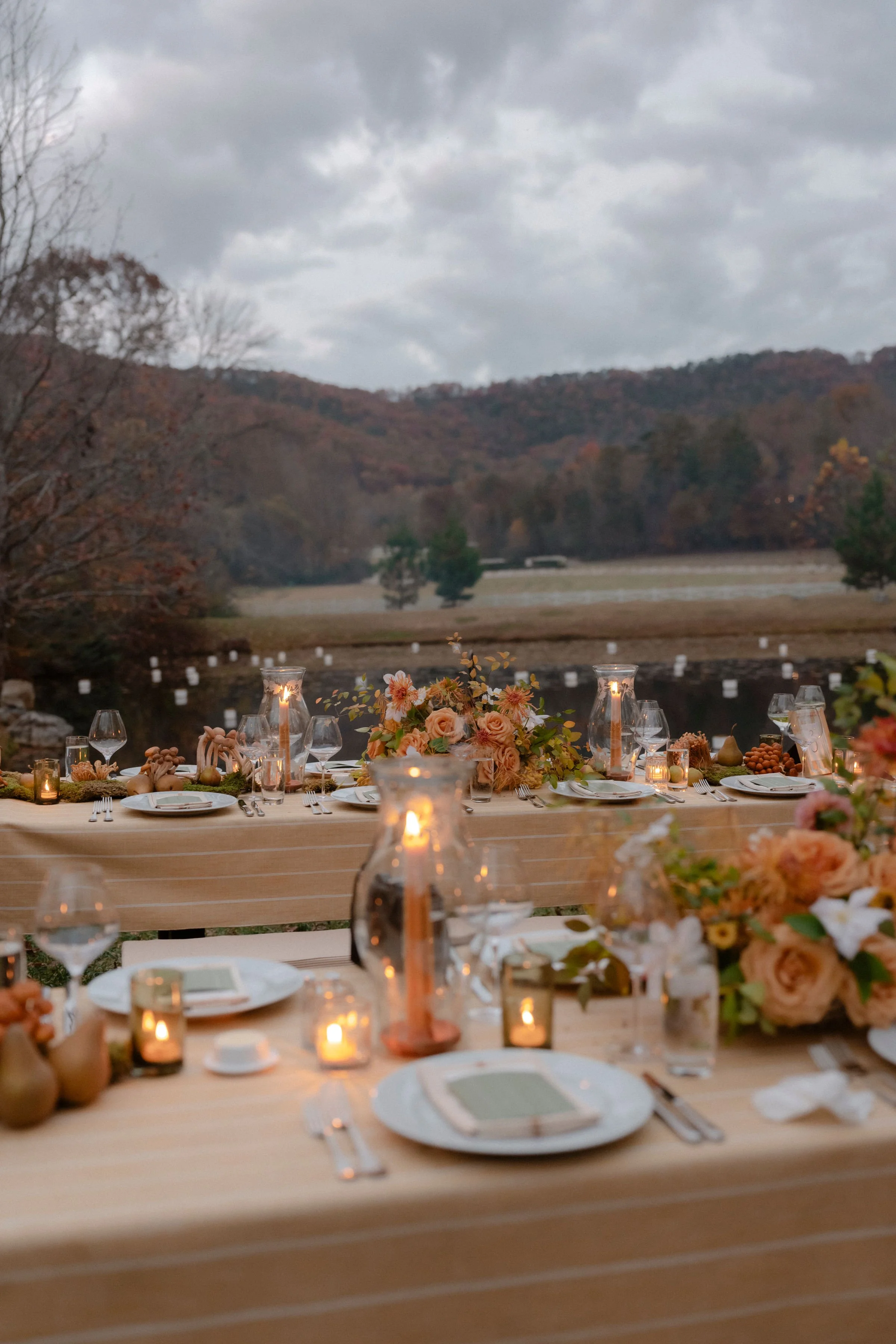 An outdoor dining setup with a long table decorated with candles, floral centerpieces, and elegant tableware. The scene is set against a backdrop of a river, trees, and rolling hills under a cloudy sky.