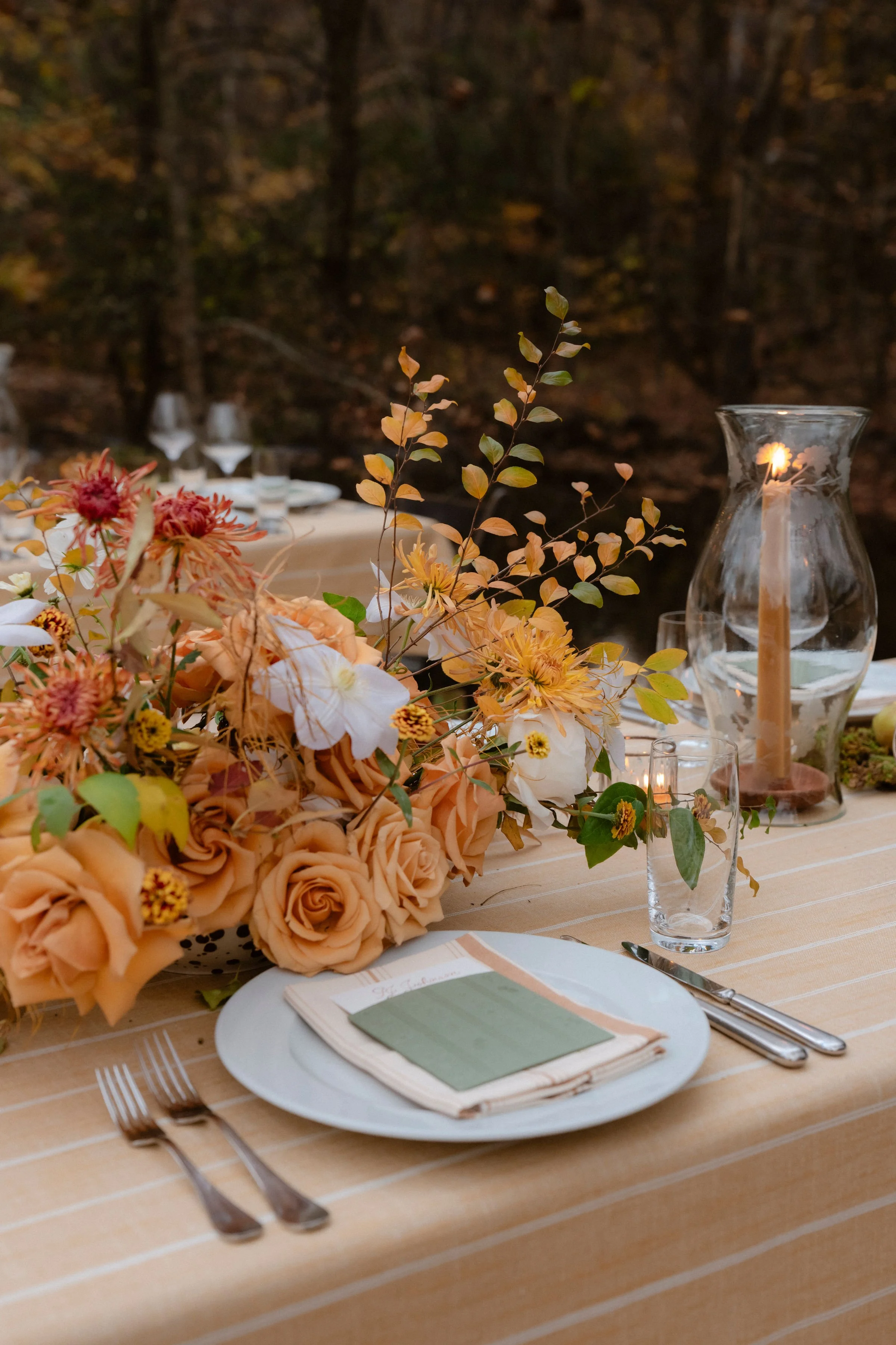 Elegant outdoor table setting with a floral centerpiece featuring peach roses, white flowers, and greenery. The table has a beige striped tablecloth, a white plate with a napkin and green place card, and silverware. A glass candle holder with a lit c