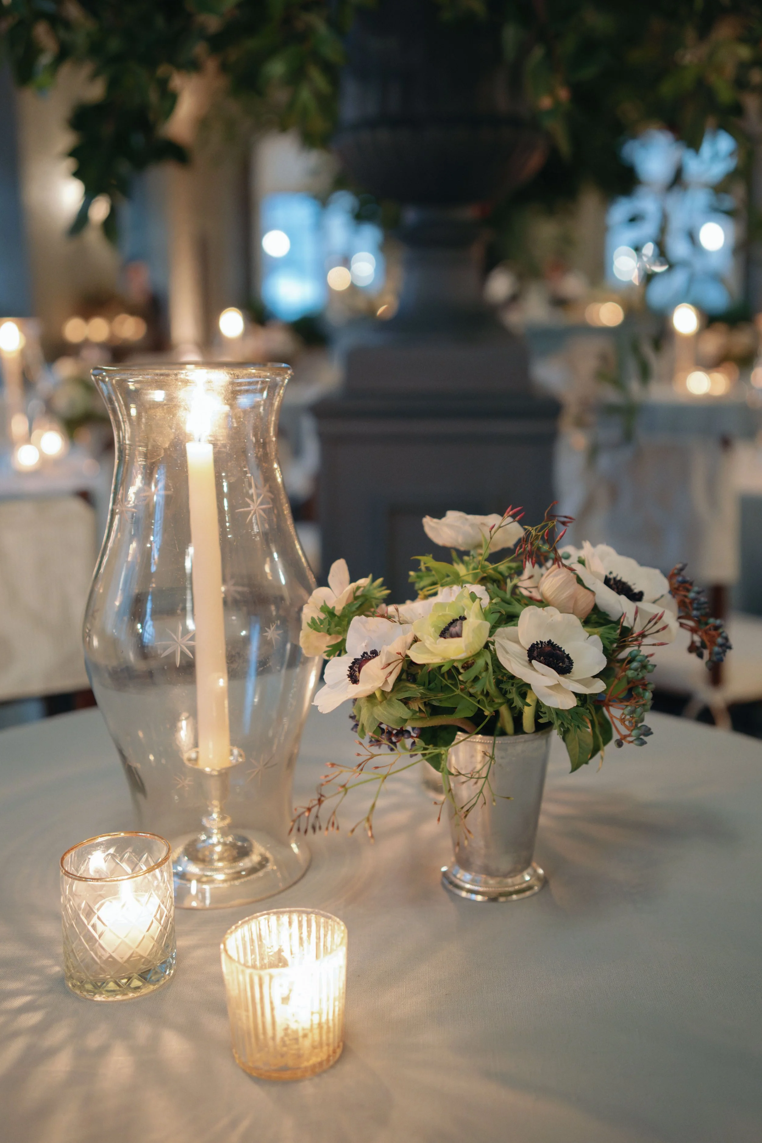 A table decorated with a large hurricane glass candle holder with a lit candle, a smaller votive candle holder with a lit candle, and a metal vase with white flowers and greenery. 