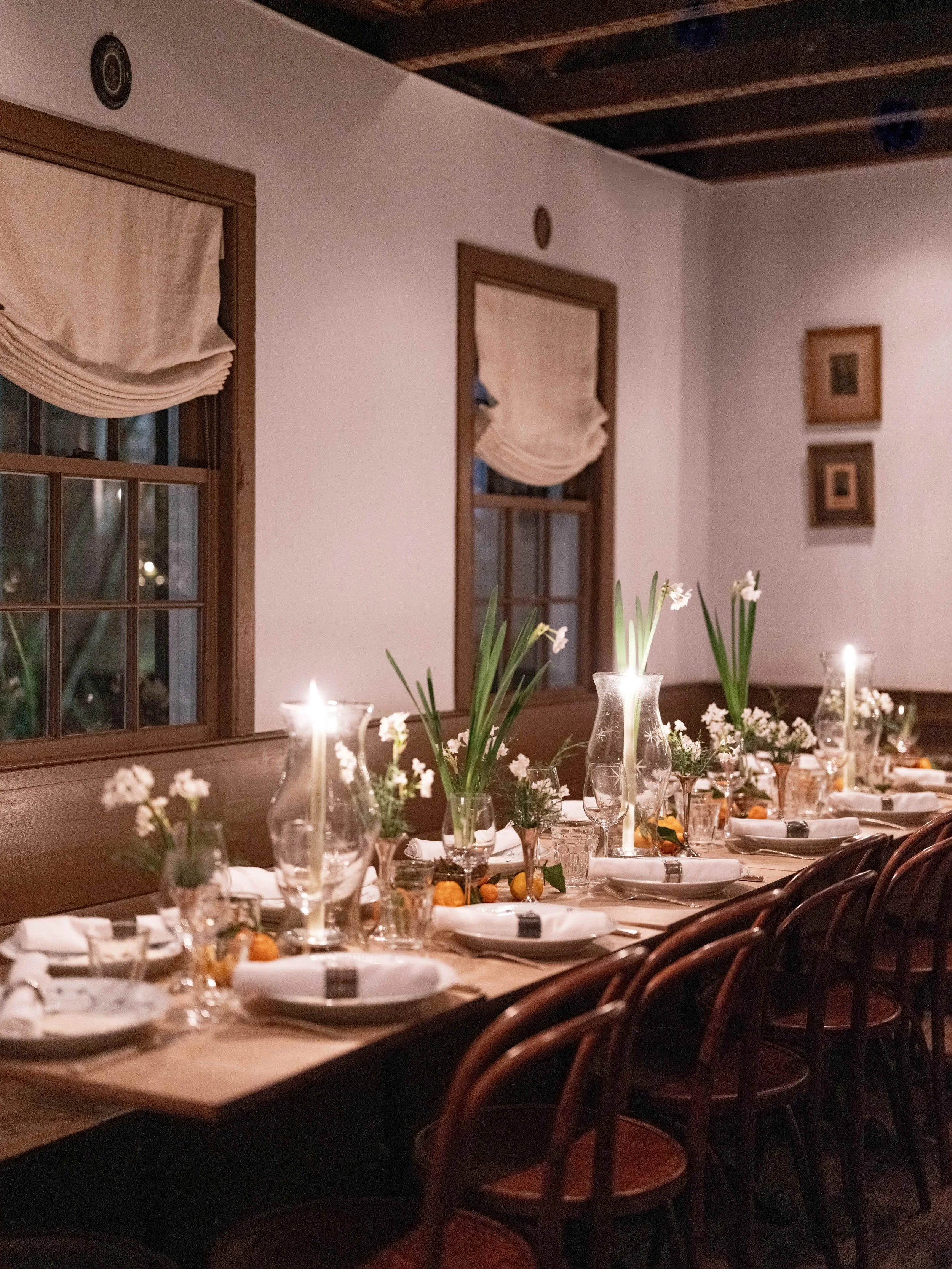 A long dining table decorated with Hurricane glass, vases with flowers, plates, glasses, and napkins in a cozy, rustic indoor setting with wooden windows and framed pictures on the wall.